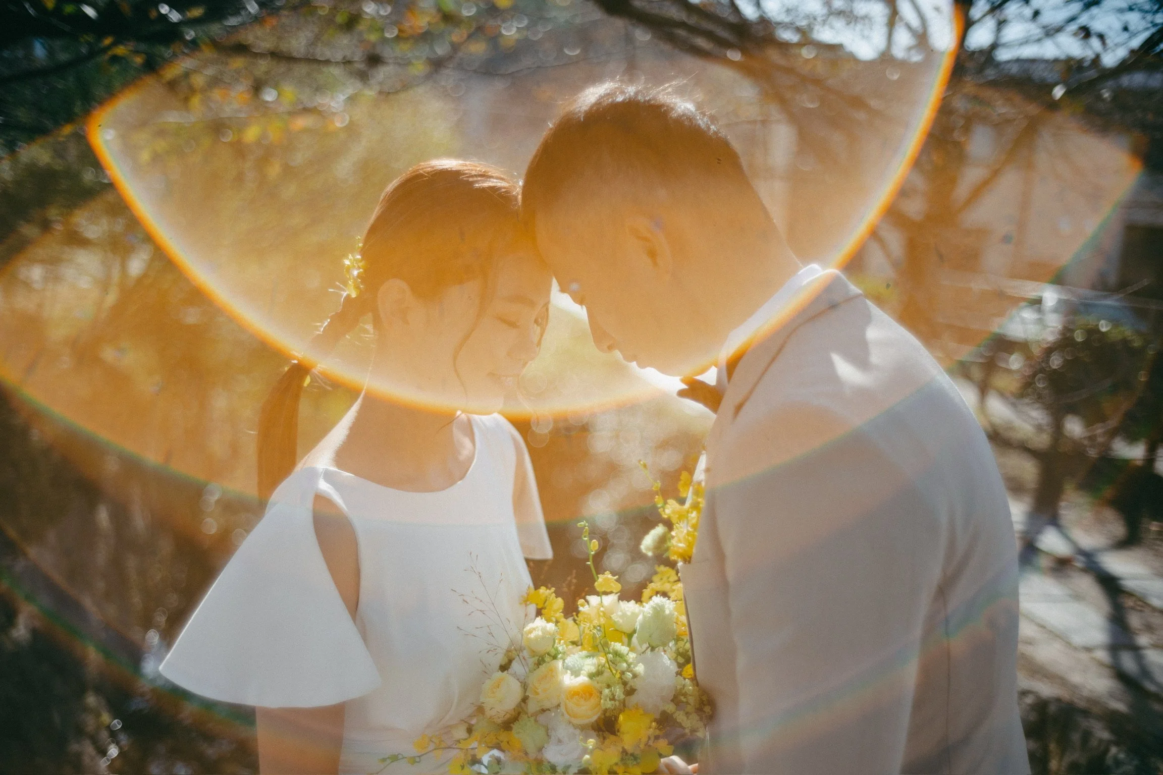 A couple on their wedding day with their foreheads touching, standing outdoors, with the bride holding a bouquet of white and yellow flowers, and sunlight creating a halo effect around them.