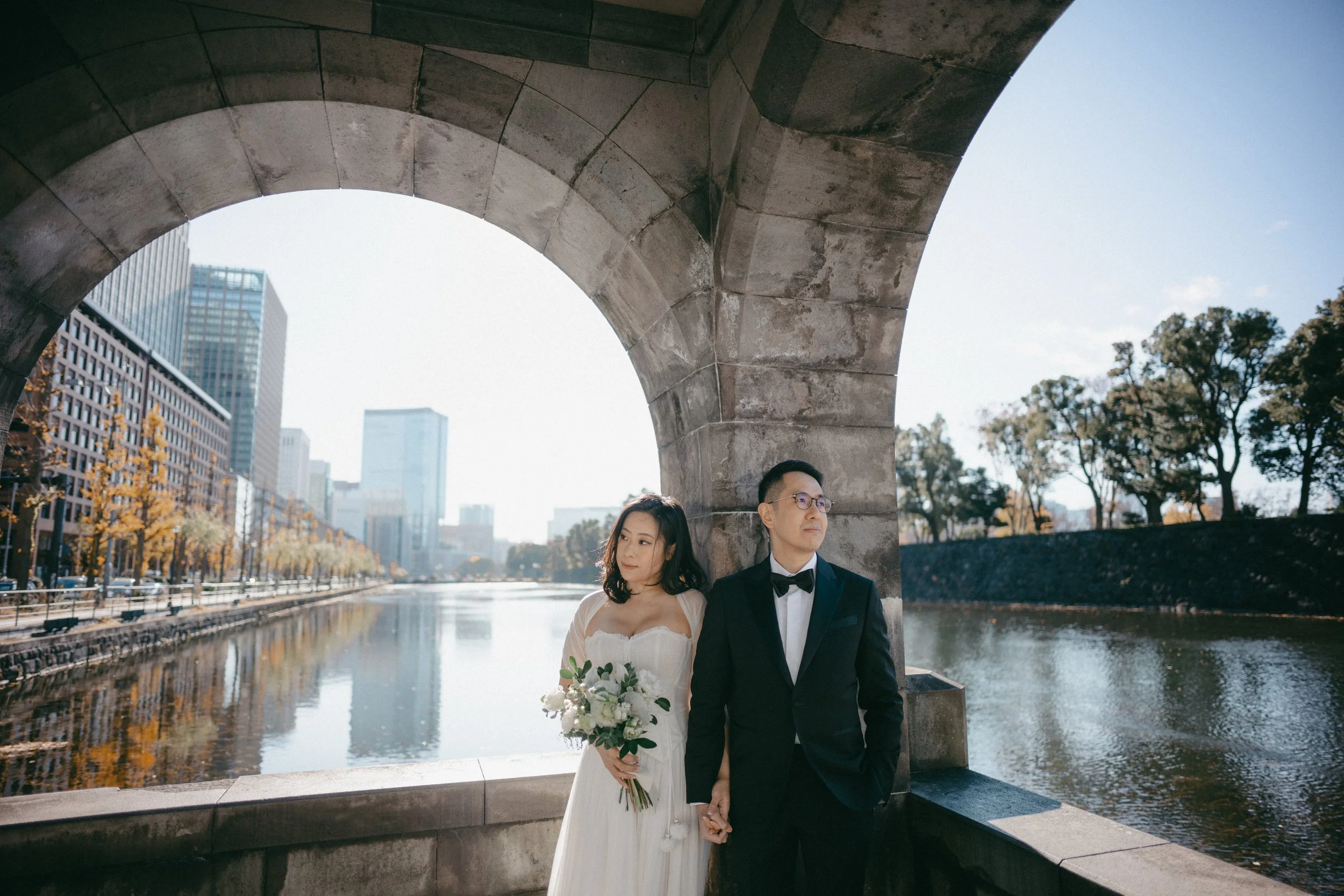 A bride and groom holding hands under a stone bridge by a river in an urban setting, with modern buildings and trees in the background on a sunny day.