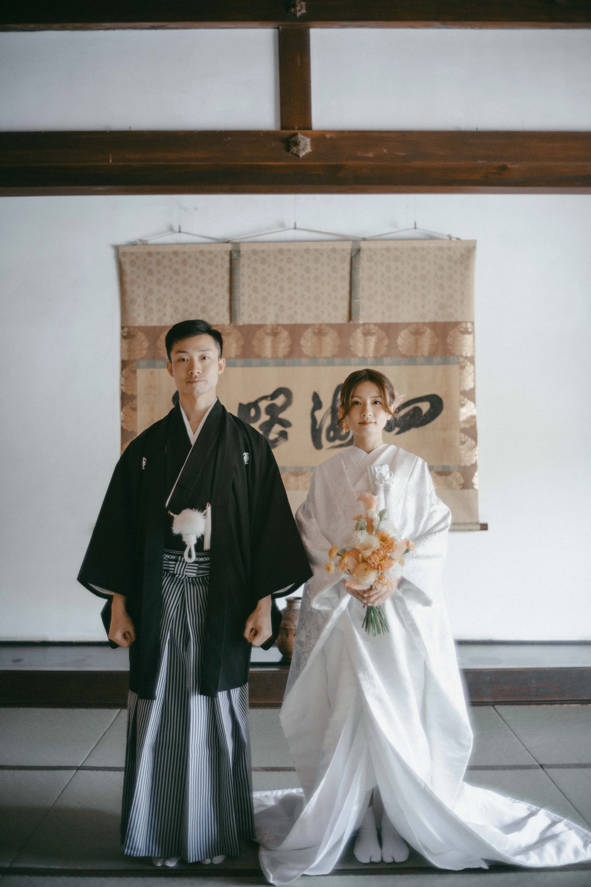 A Japanese bride and groom in traditional wedding attire standing indoors in front of a calligraphy scroll.