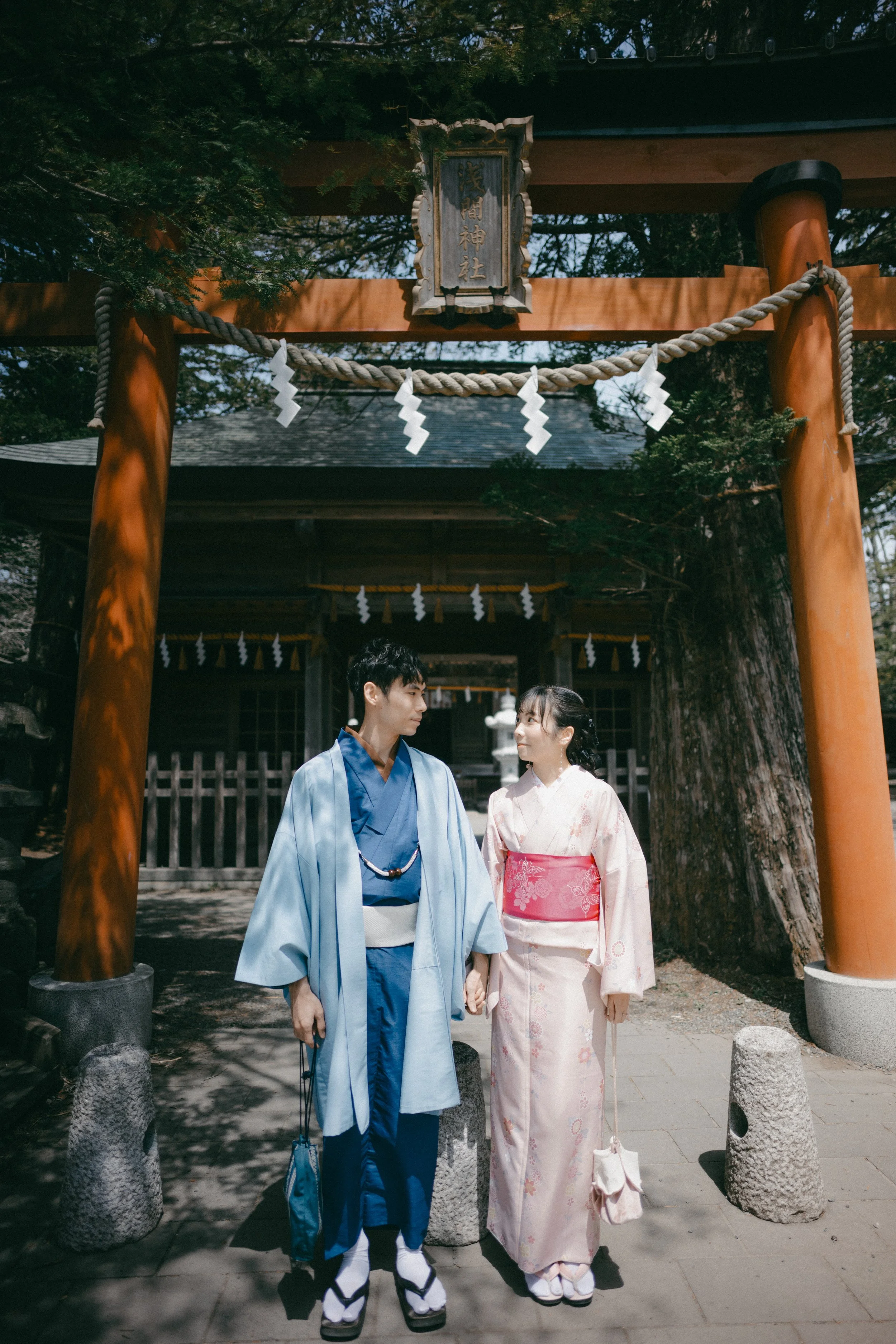 A young couple in traditional Japanese kimonos standing hand in hand in front of a Shinto shrine entrance with a torii gate, shide paper streamers, and a large tree in the background.
