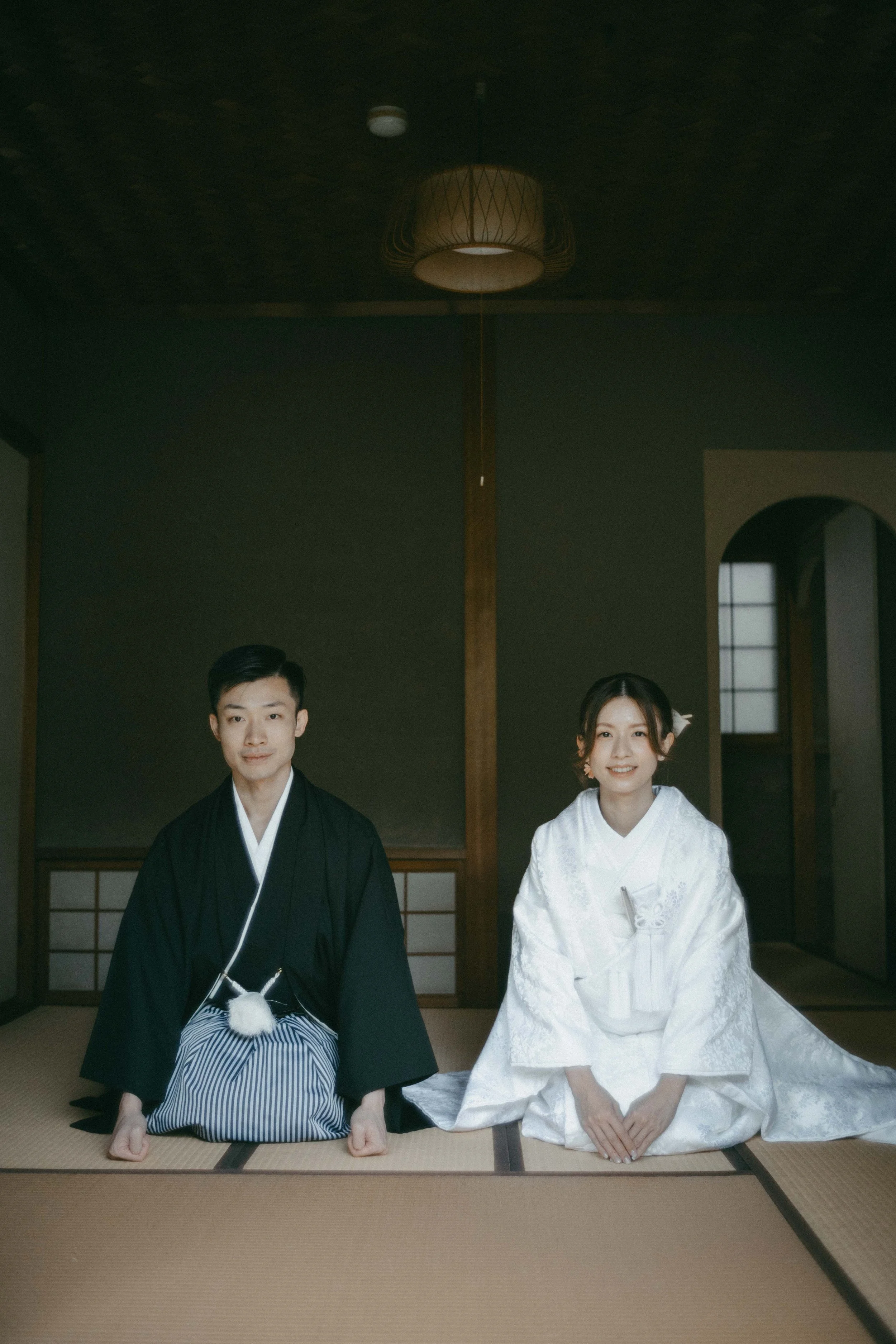 A bride and groom dressed in traditional Japanese wedding attire seated on tatami mats inside a room with shoji screens and wooden accents.