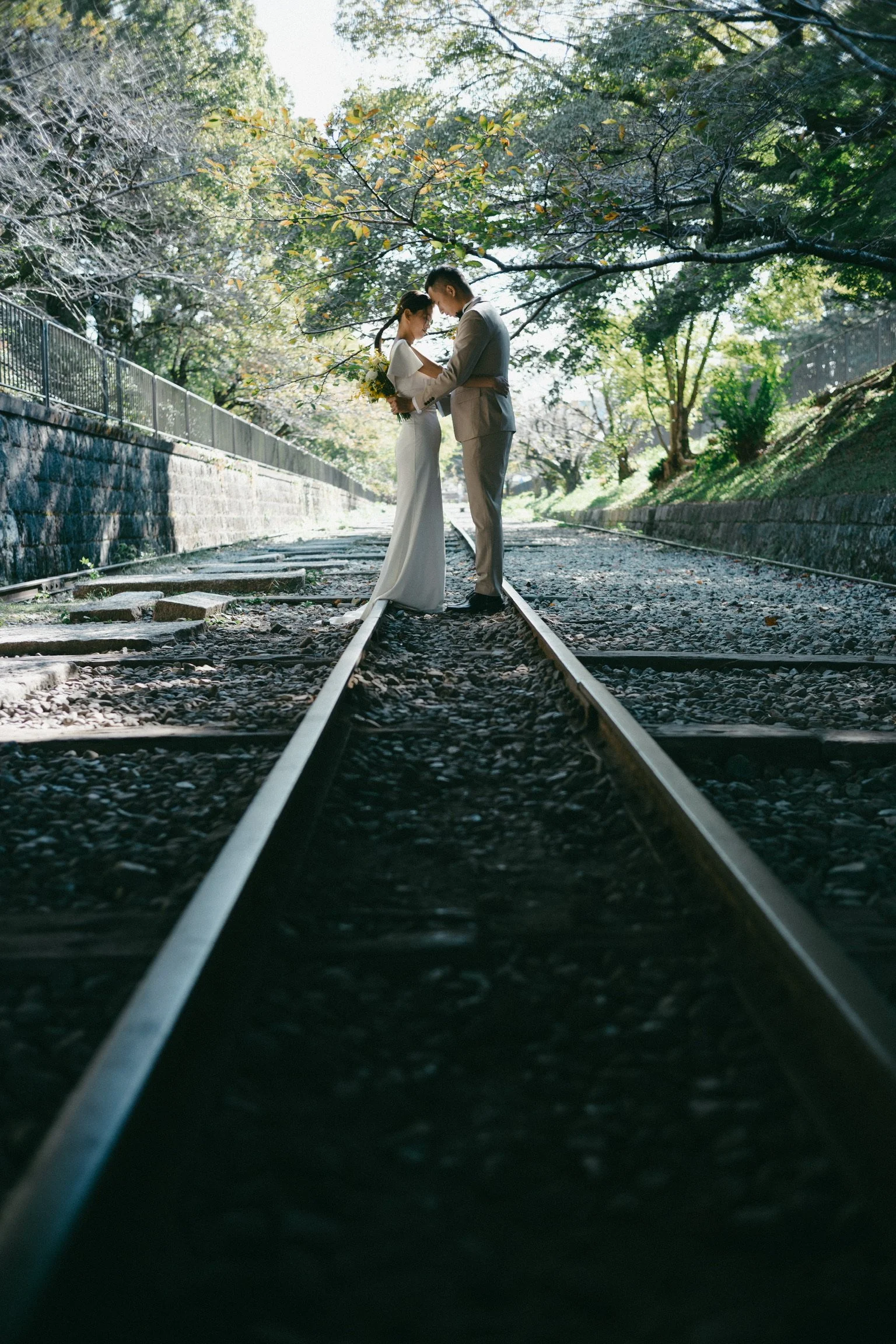 A bride and groom standing together on railway tracks in a green, tree-filled outdoor setting, holding hands and looking at each other. The bride is wearing a white wedding dress and the groom a gray suit.