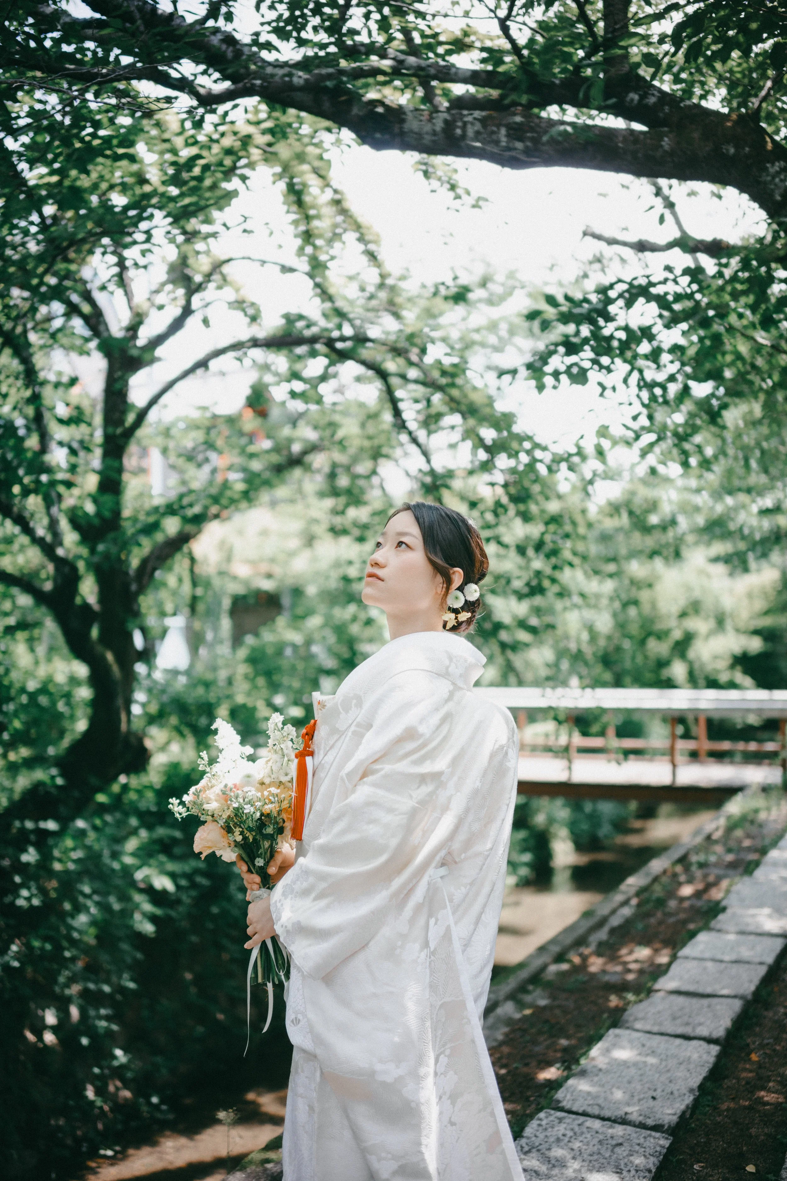 A woman dressed in a traditional white kimono with floral details, holding a bouquet of flowers, standing outdoors near a stone wall and a small bridge, surrounded by green trees.