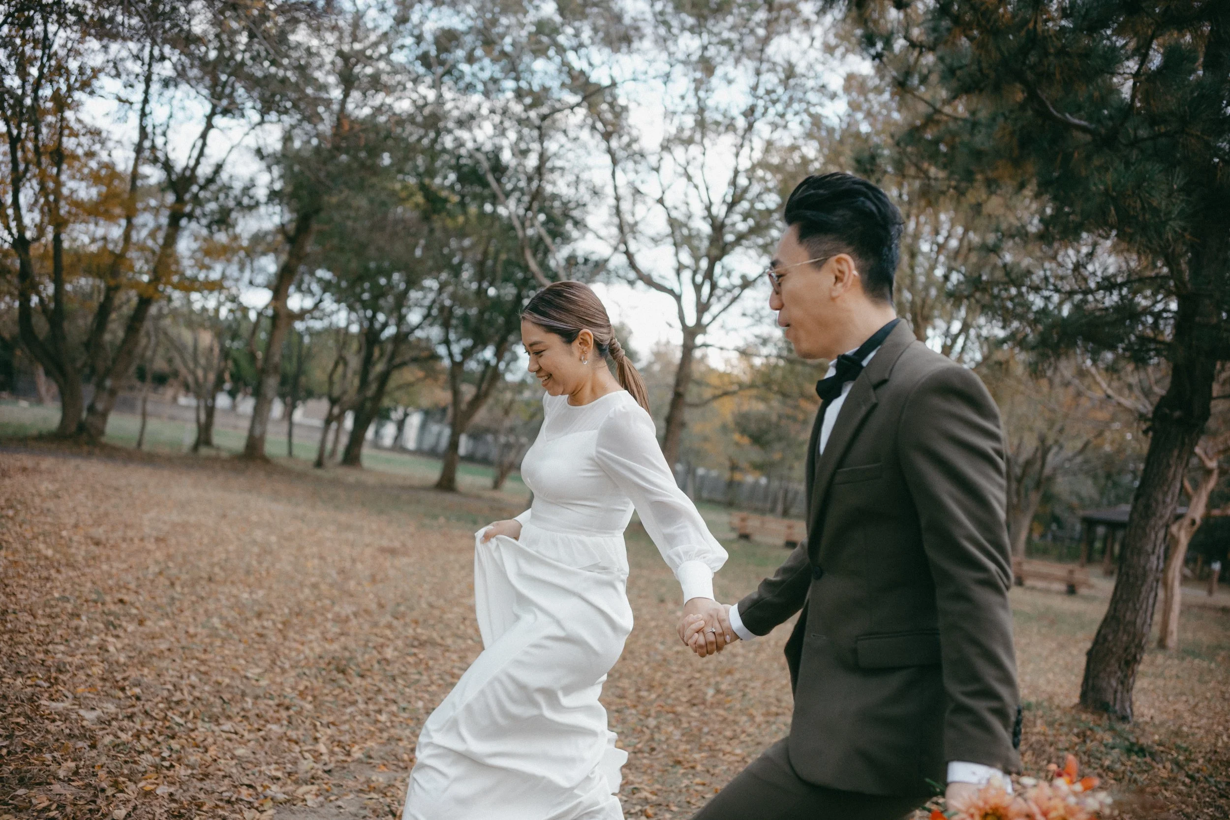 A couple walking hand in hand through a park with fall leaves on the ground and trees in autumn colors, smiling and enjoying each other's company.