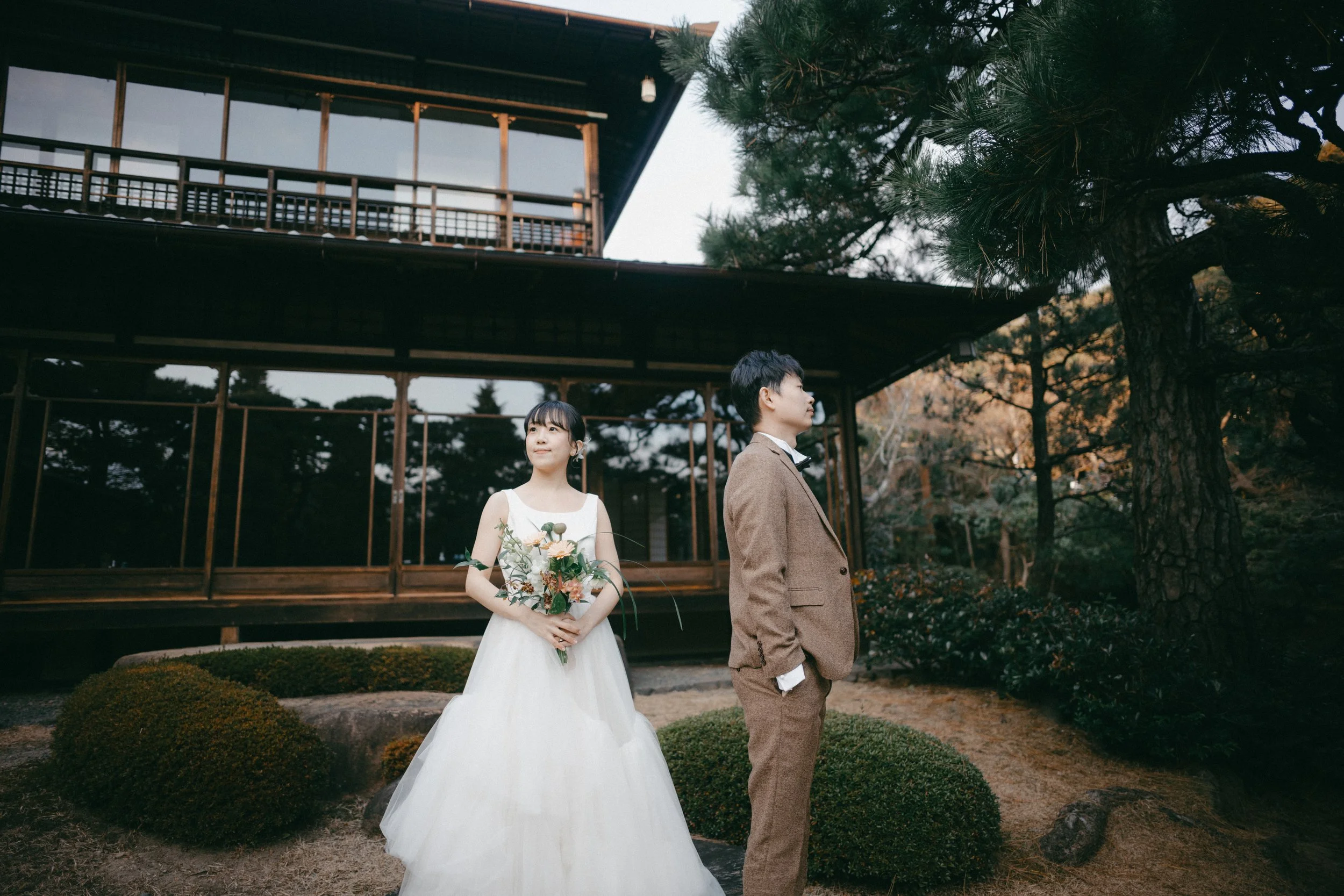 A bride in a white wedding dress holding a bouquet and a groom in a brown suit standing outdoors in front of a traditional Japanese wooden building surrounded by trees.