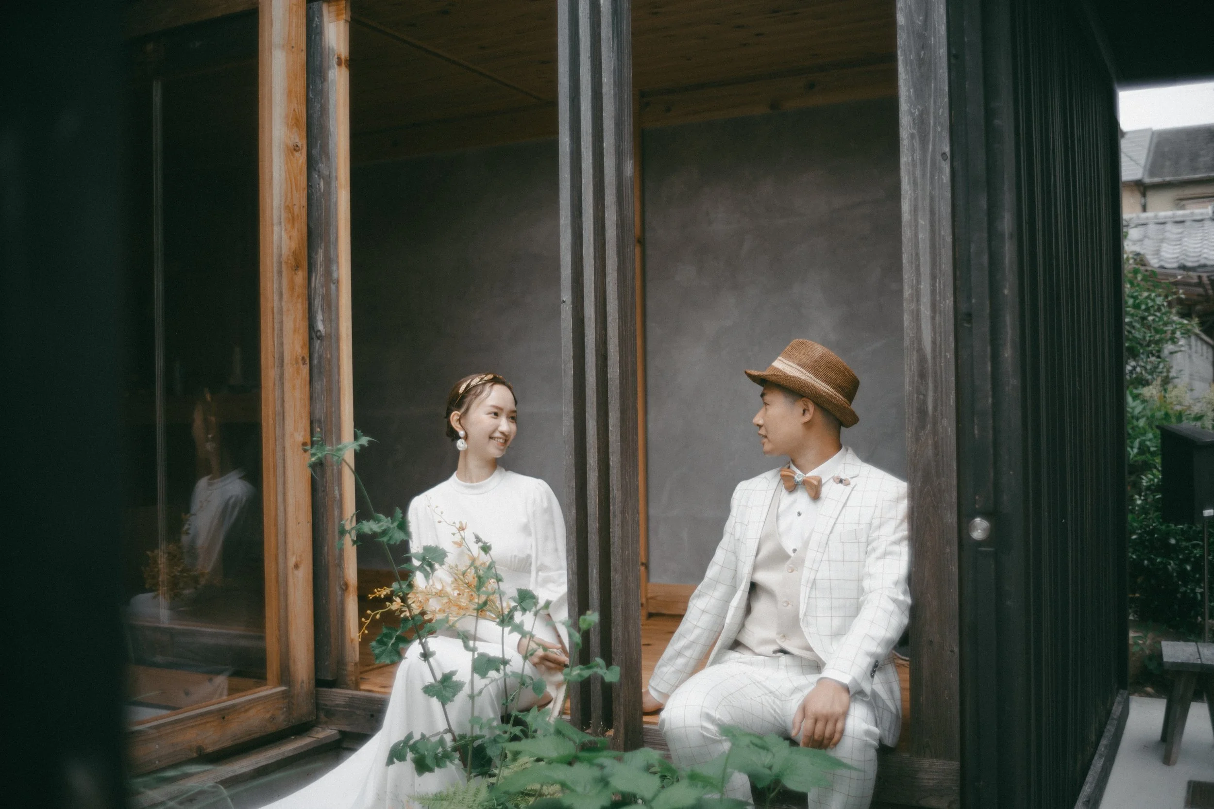 A man and woman dressed in vintage white attire sitting on a wooden platform, smiling at each other beside a glass window with greenery outside, in a modern rustic setting.