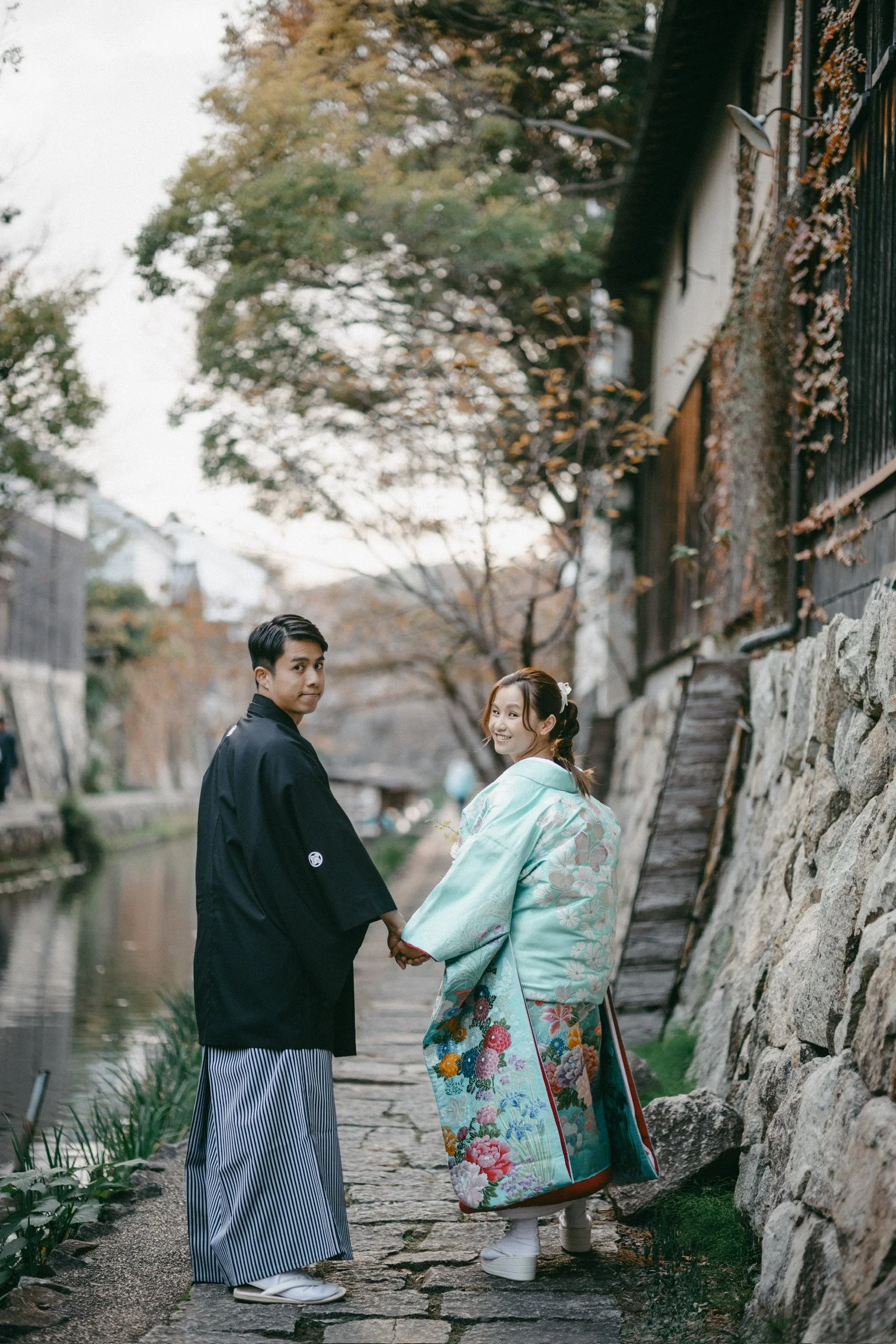 A young couple dressed in traditional Japanese attire holding hands and walking along a stone path beside a canal, with autumn trees and old buildings in the background.