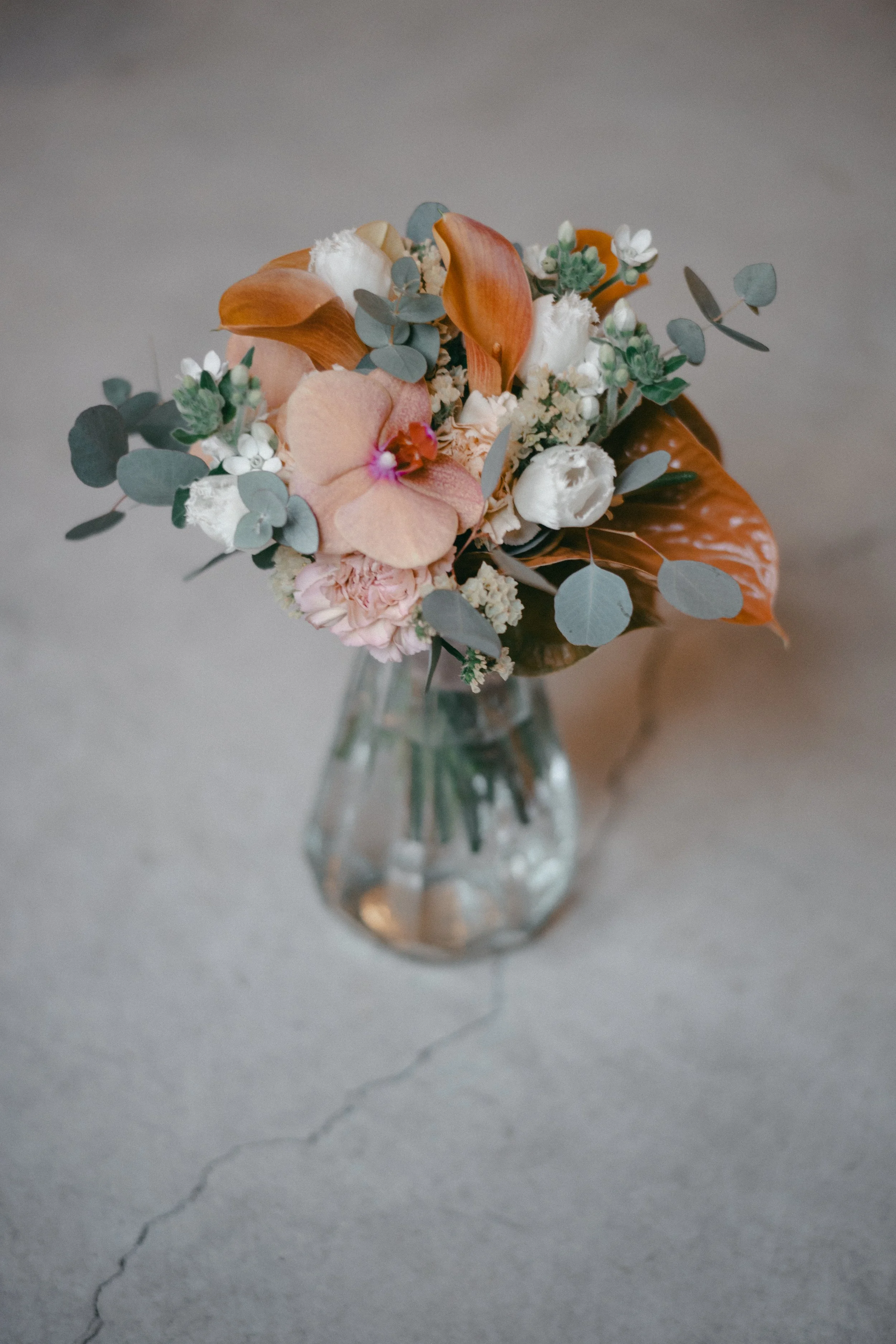 A flower arrangement in a clear glass vase with pink, white, and orange flowers and green foliage on a concrete surface.