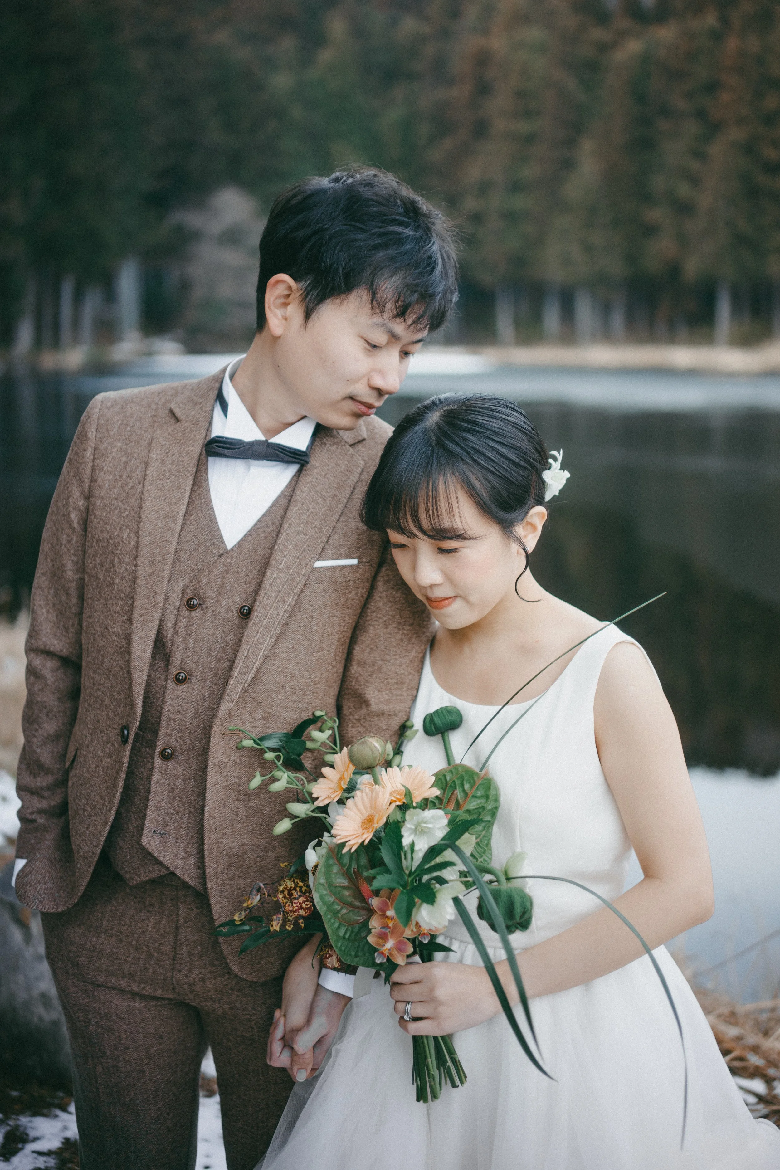 A couple dressed in wedding attire, the man in a brown suit and the woman in a white dress, holding hands near a lake with a forest in the background, the woman holding a bouquet of flowers.