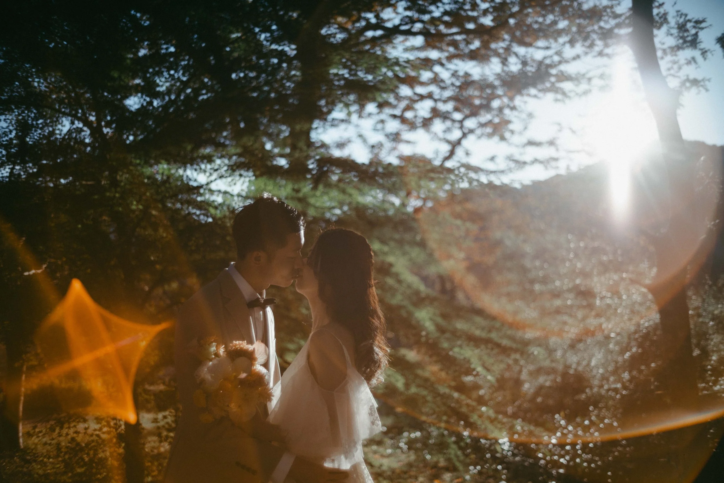 A couple in wedding attire sharing a kiss outdoors at sunset with trees and sunlight in the background.