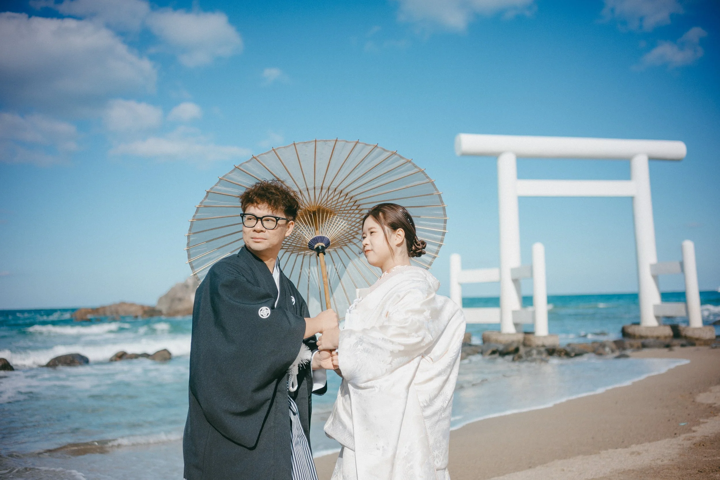 A couple in traditional Japanese wedding attire standing on a beach, holding an umbrella, with a torii gate and ocean in the background.