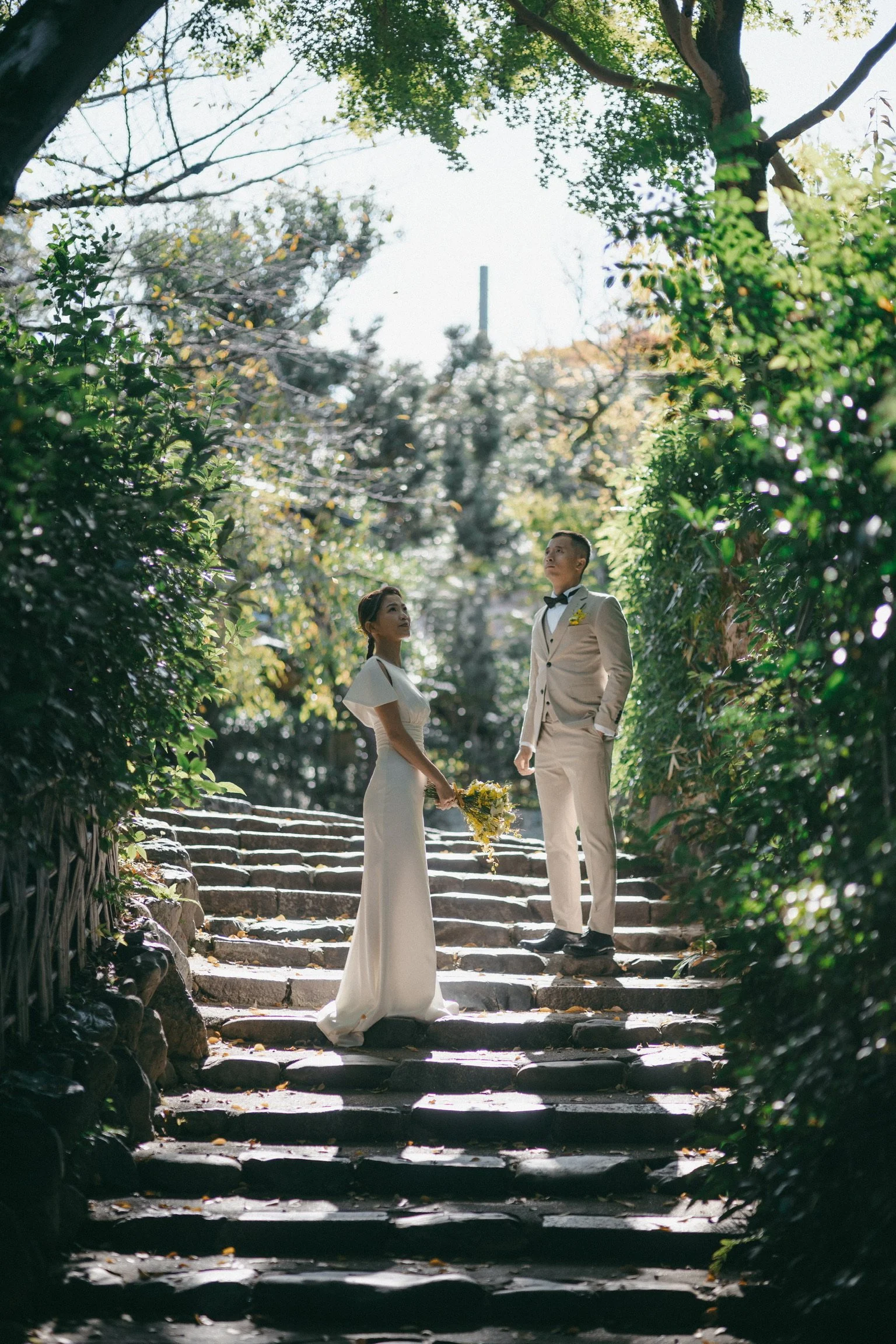 A bride and groom stand on stone steps in a lush garden, gazing at each other. The bride holds a bouquet of yellow flowers, and both are dressed in wedding attire with sunlight filtering through trees.