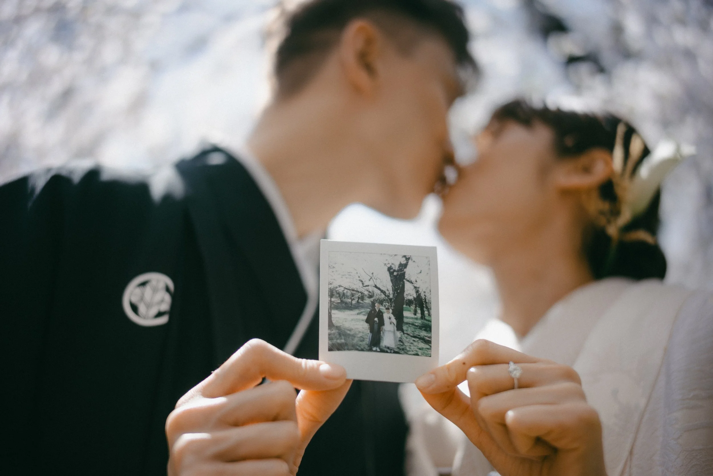 A couple holding a black and white photo of themselves with a tree in the background, as they share a kiss.