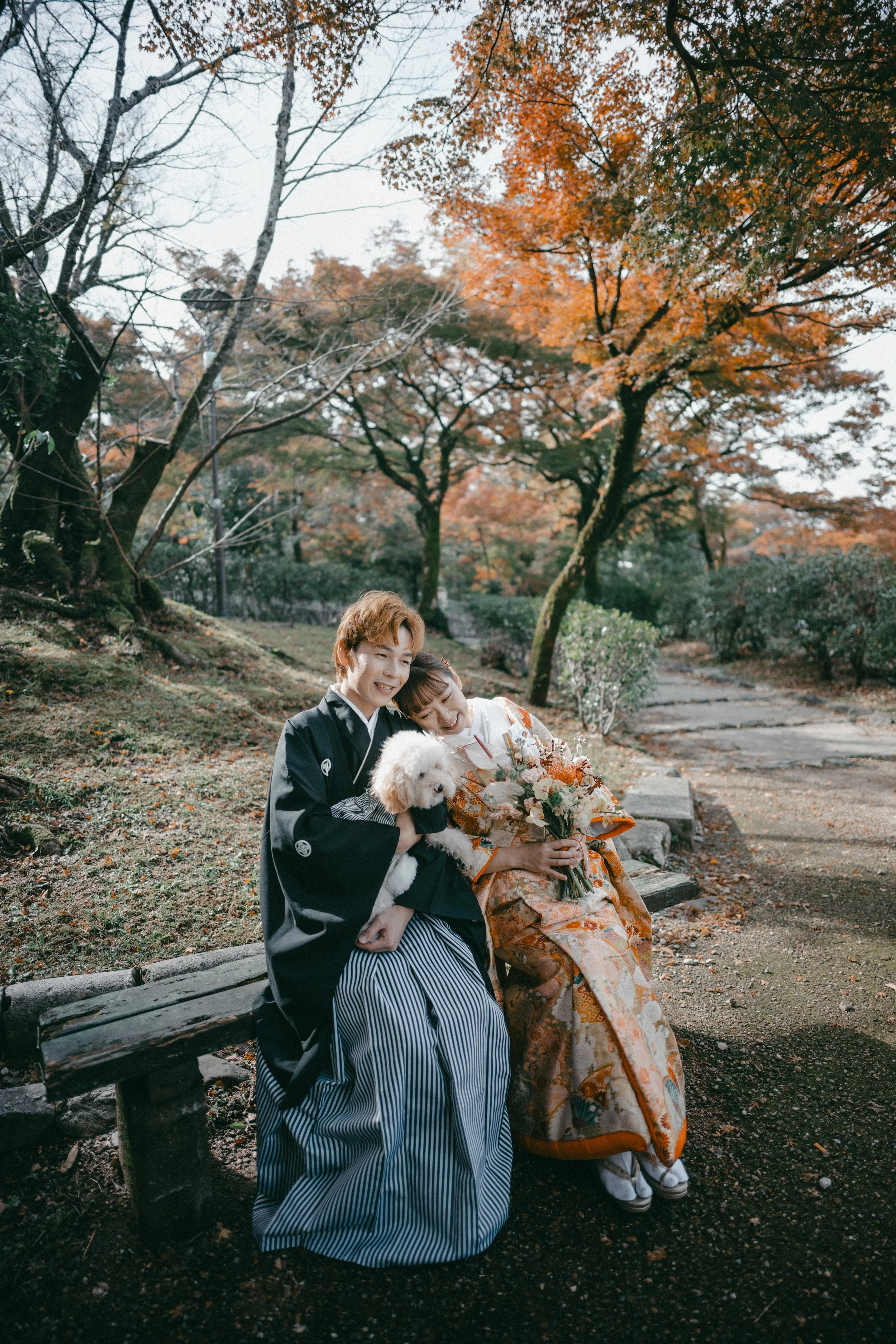 Two women in traditional Japanese kimonos sitting on a park bench in autumn, smiling and cuddling a small white dog, with orange and green fall foliage surrounding them.