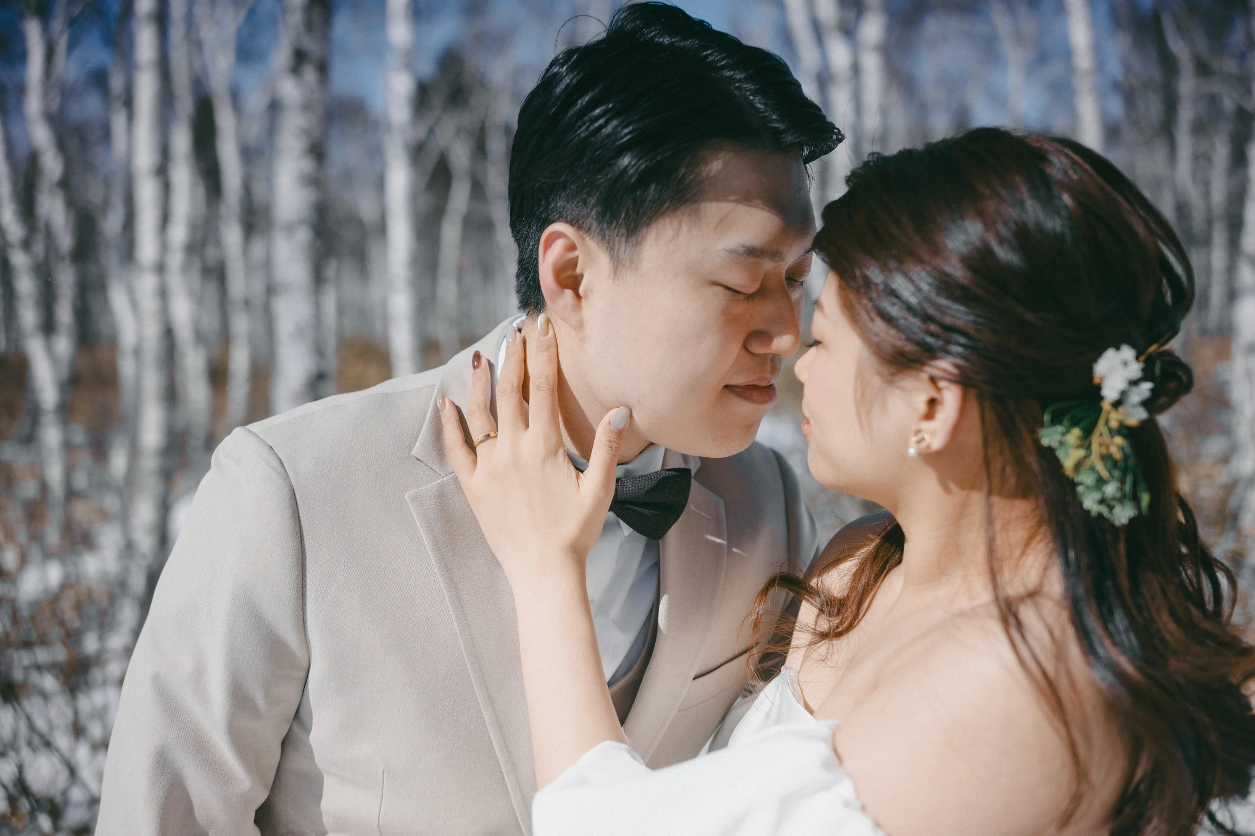 A couple in wedding attire, the man in a light-colored suit with a black bow tie, and the woman with long dark hair and a floral hairpiece, stand close with foreheads touching in a snowy forest.