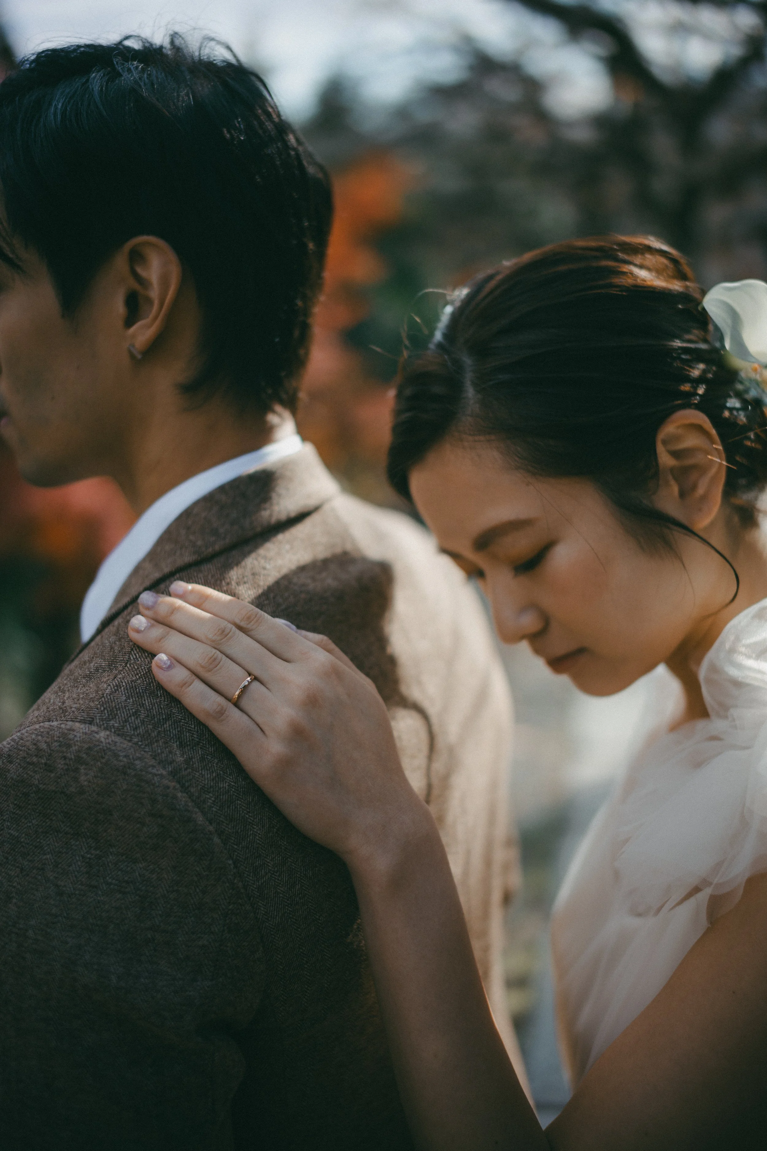 A woman gently resting her hand on a man's shoulder, both with their eyes closed, outdoors with autumn trees in the background.