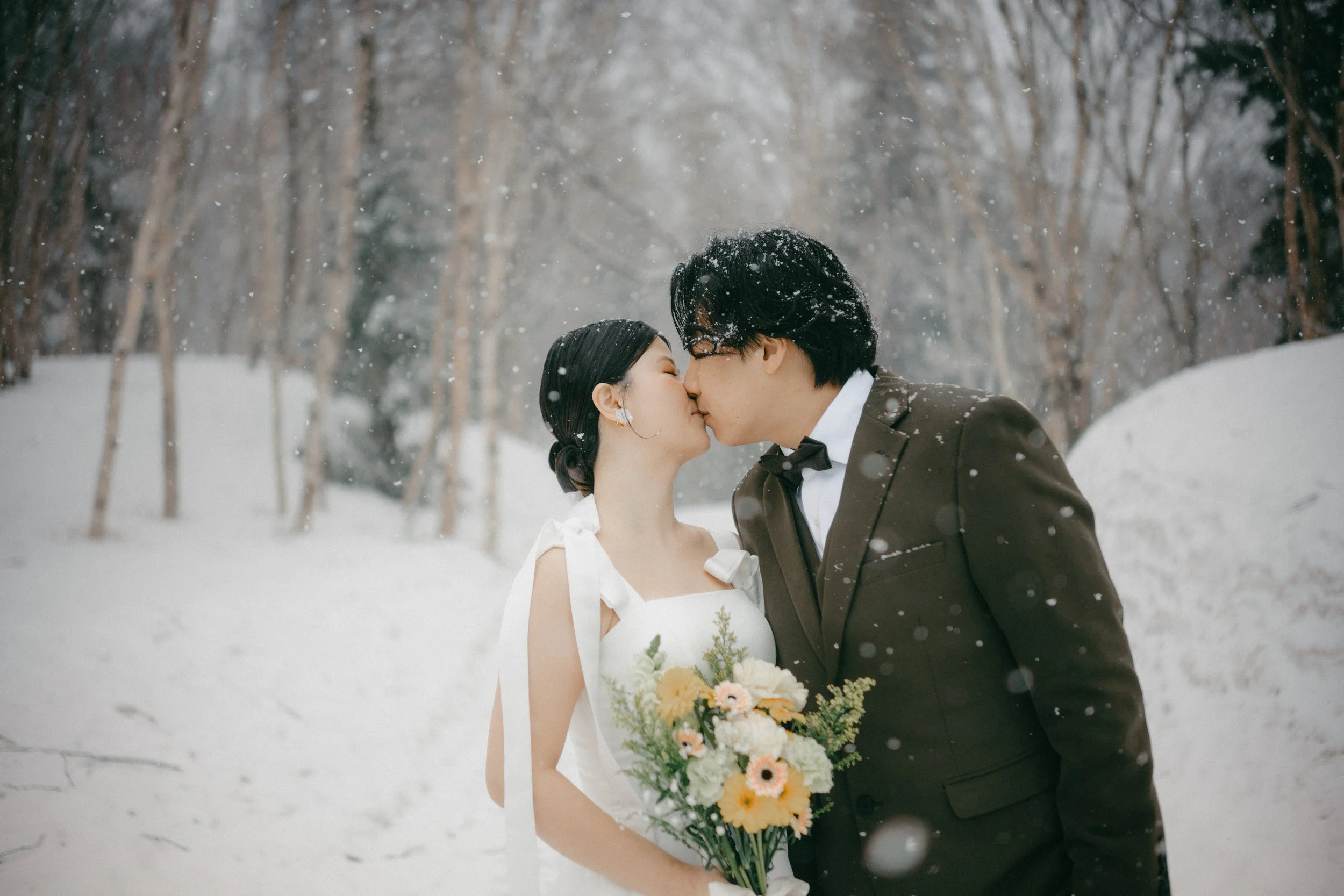 A bride and groom kissing in a snowy forest, the bride holding a bouquet of yellow and white flowers.