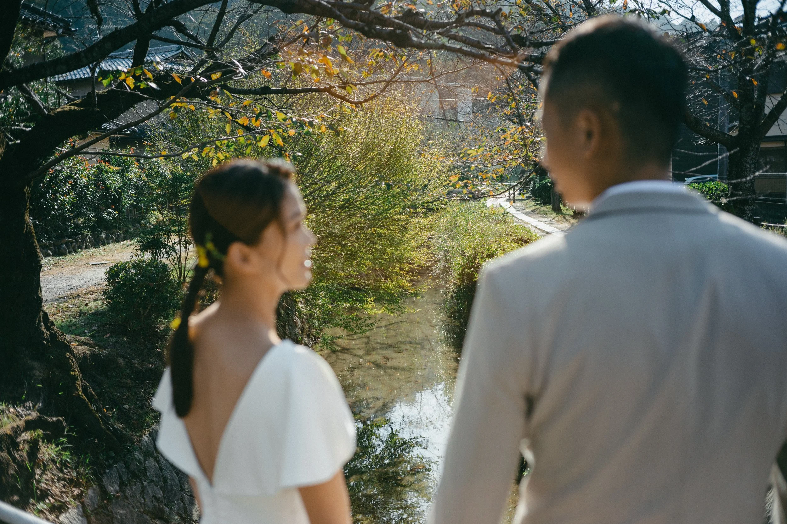 A man and woman standing outdoors near a small stream, with trees and greenery around, on a sunny day. They are smiling and facing each other.