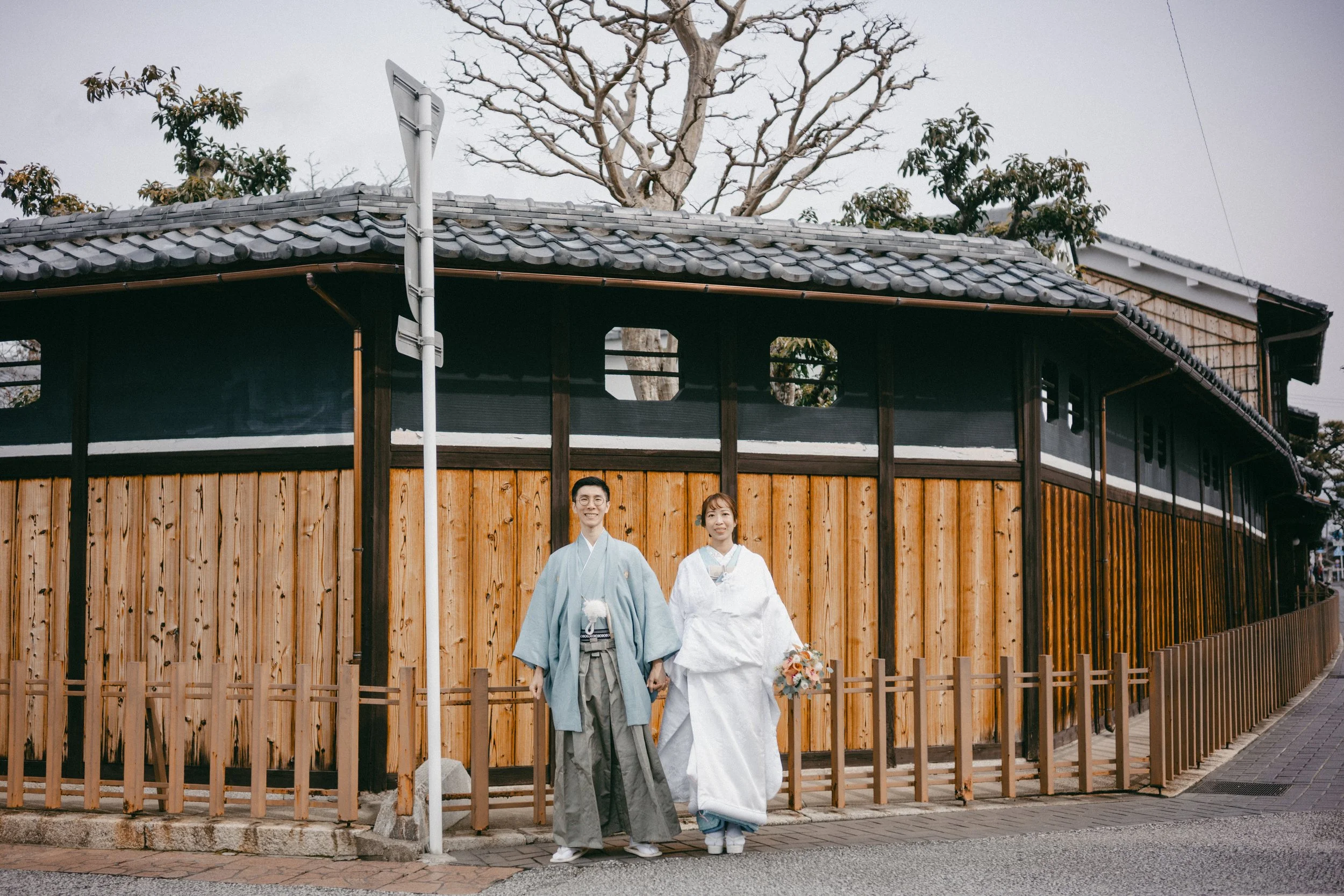 A couple dressed in traditional Japanese wedding attire standing in front of a wooden building with a tiled roof.