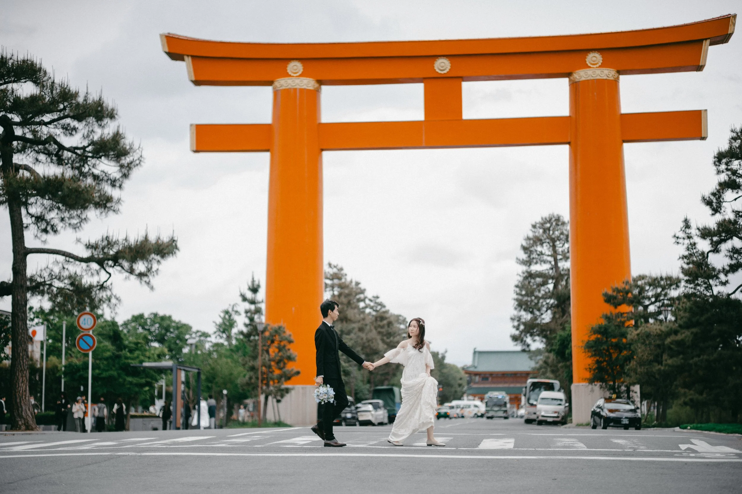 A couple walking hand in hand across a crosswalk in front of a large red torii gate, with trees and cars in the background.