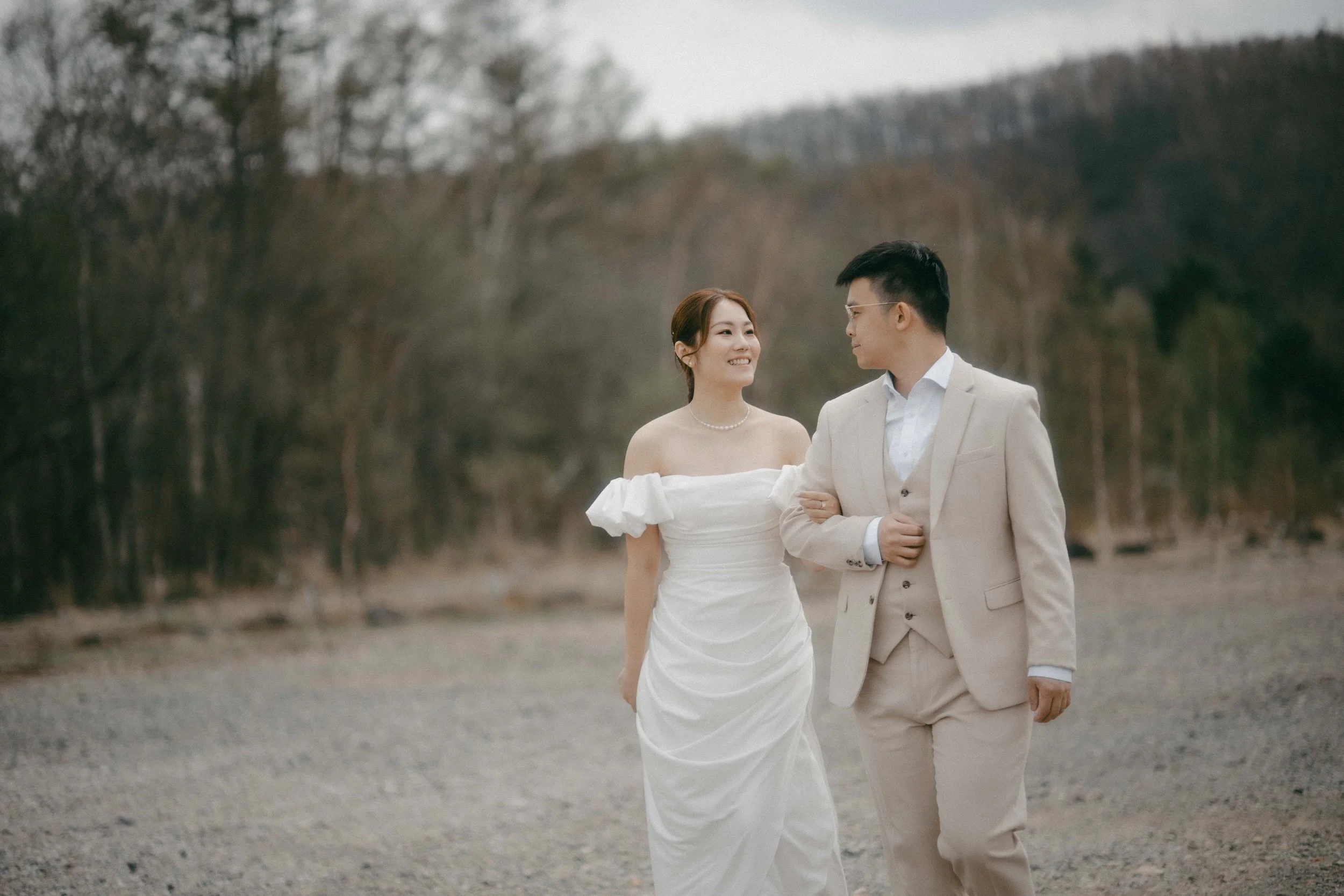 A bride and groom walking outdoors, holding arms, with trees and hills in the background.