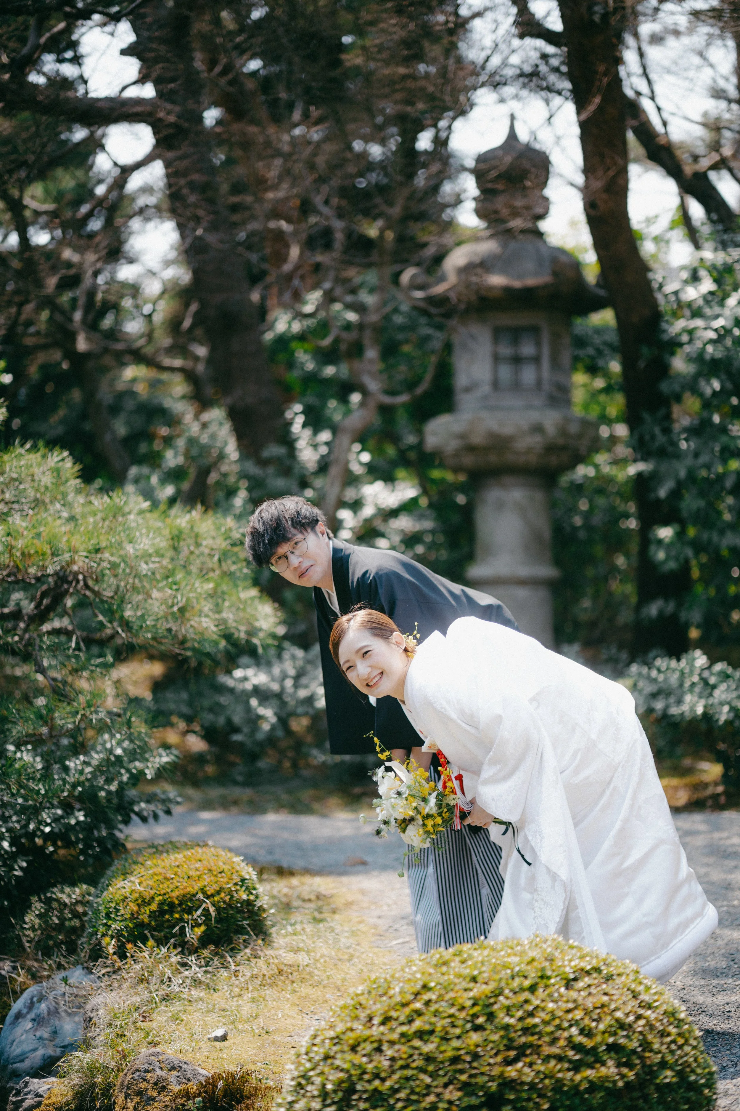 A Japanese bride and groom bowing and smiling during their wedding ceremony in a traditional garden, with greenery, rocks, and a stone lantern in the background.