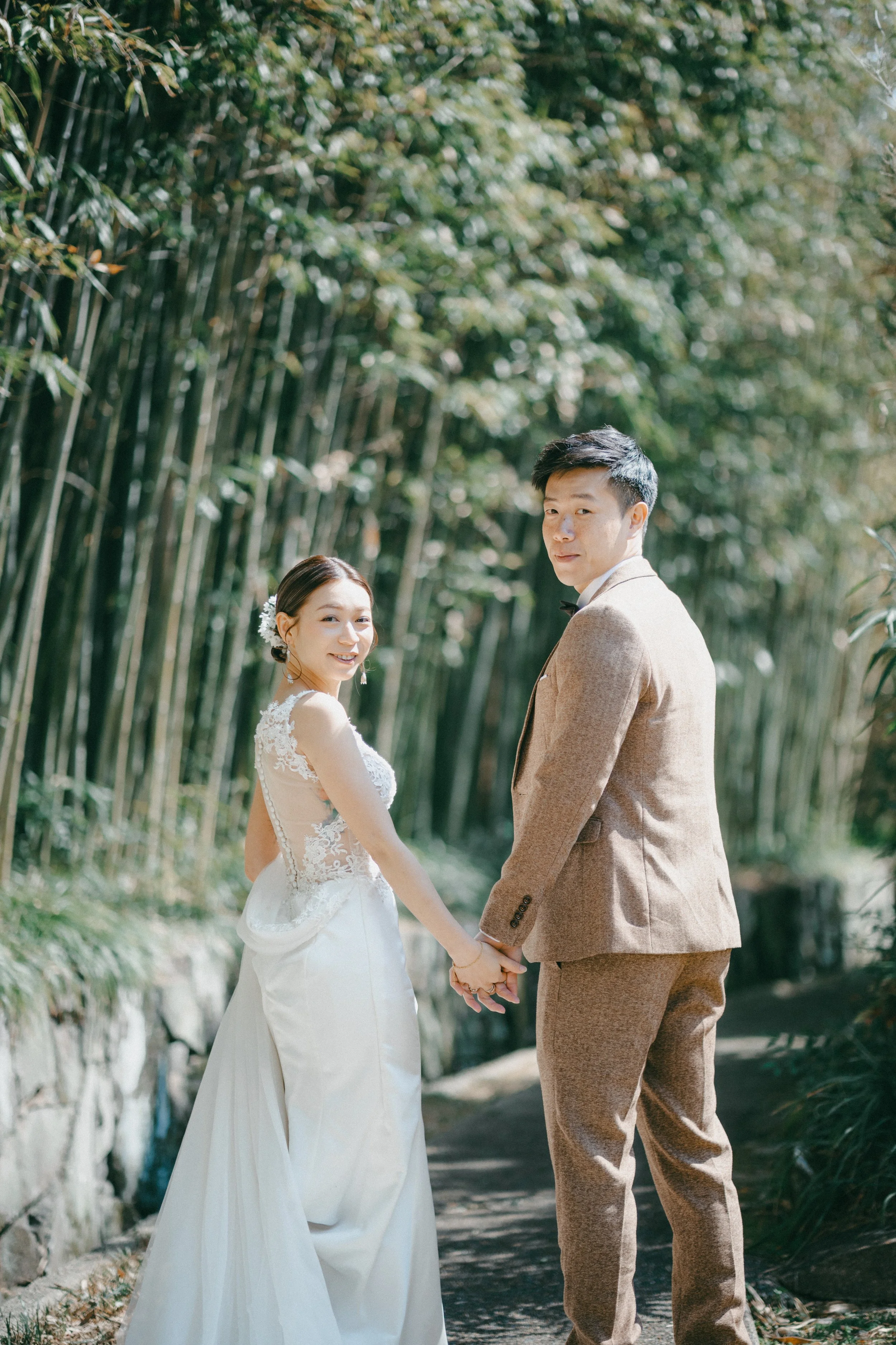A bride and groom holding hands and smiling in a lush bamboo forest.