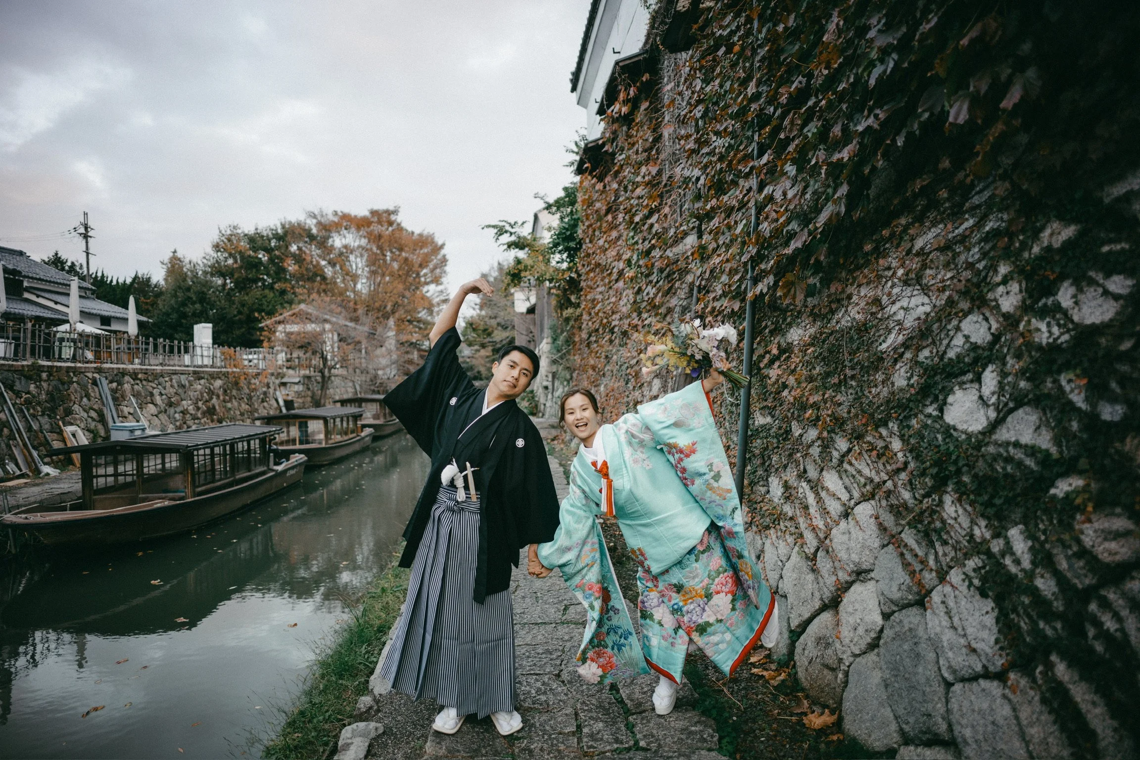 A young couple dressed in traditional Japanese kimonos and hakama posing playfully on a stone pathway by a canal, with wooden boats and a stone wall covered in ivy behind them.