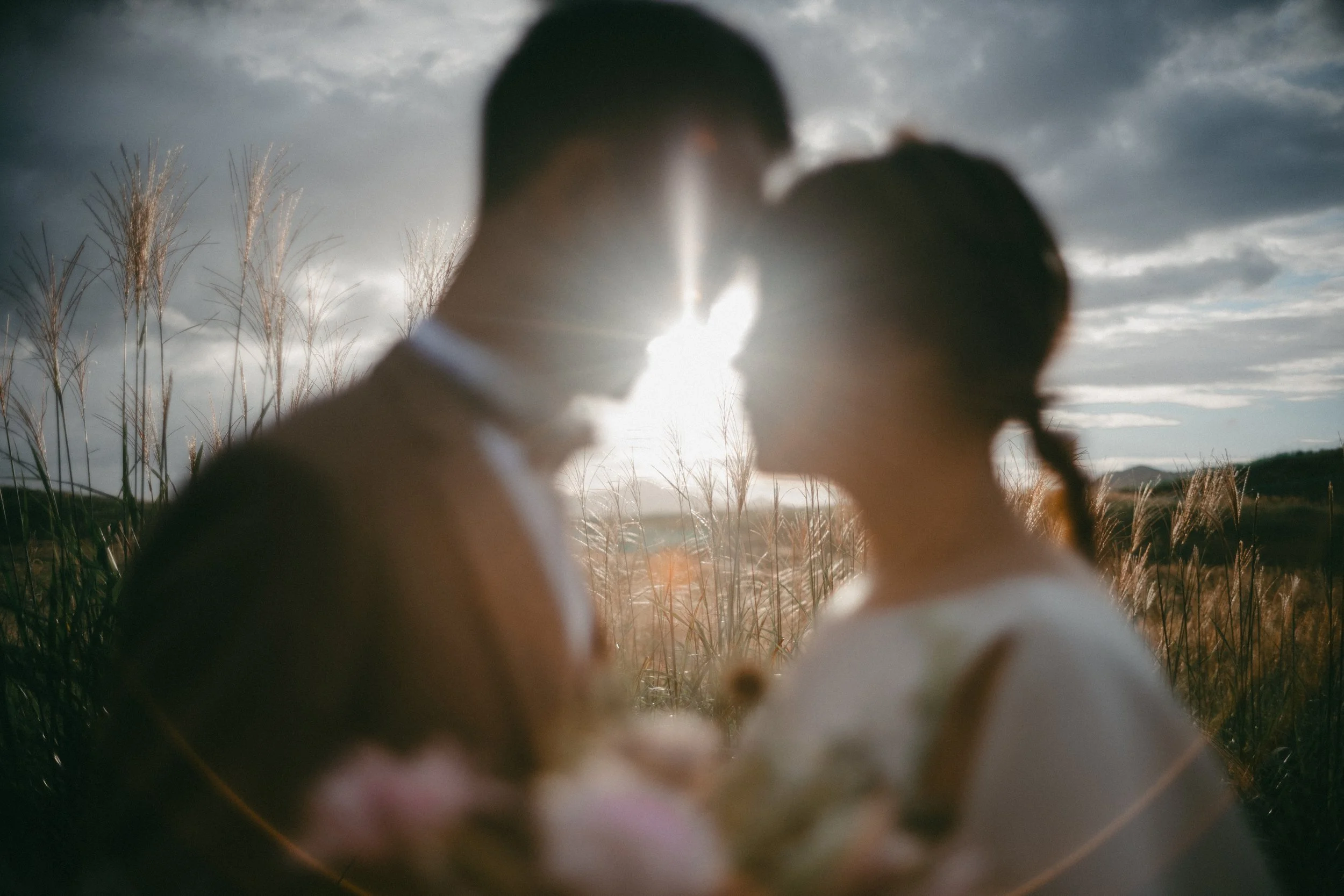 A couple is standing close together outdoors, with the sun setting behind them, creating a backlit effect. Their faces are near each other, suggesting intimacy, with a field of tall grasses in the background.