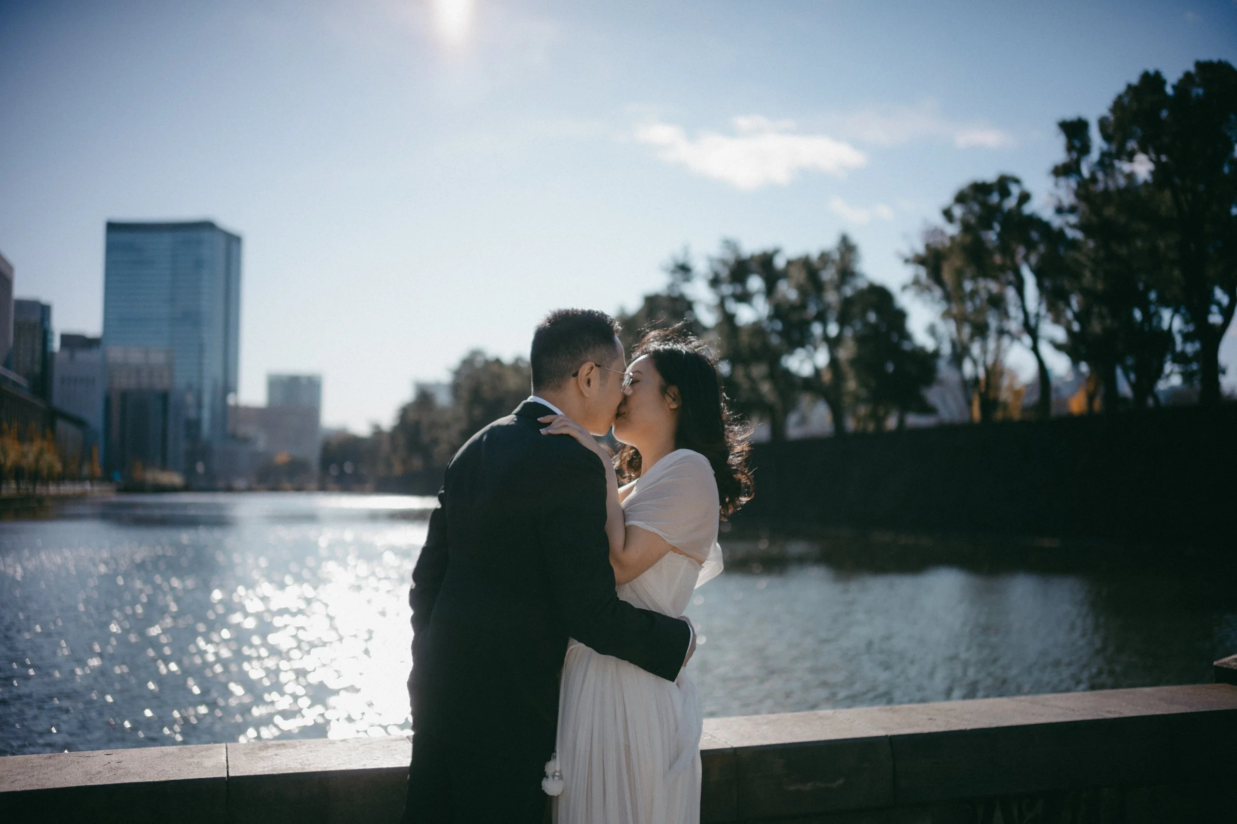 A couple kissing by a river in an urban setting with trees and city buildings in the background.