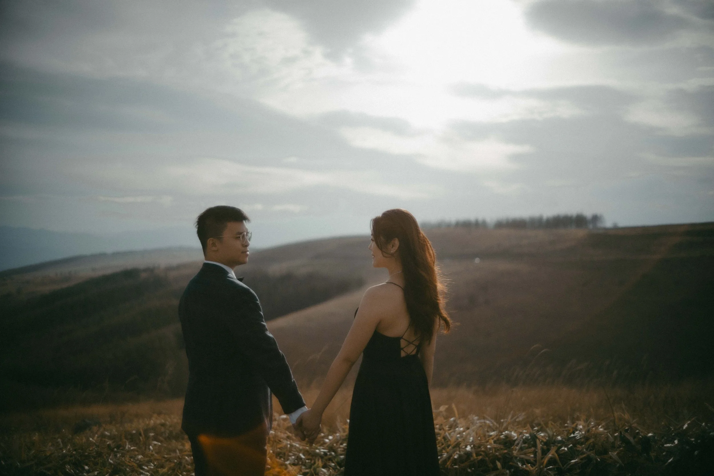 A man and a woman holding hands in a field with rolling hills in the background, looking at each other during sunset or sunrise.