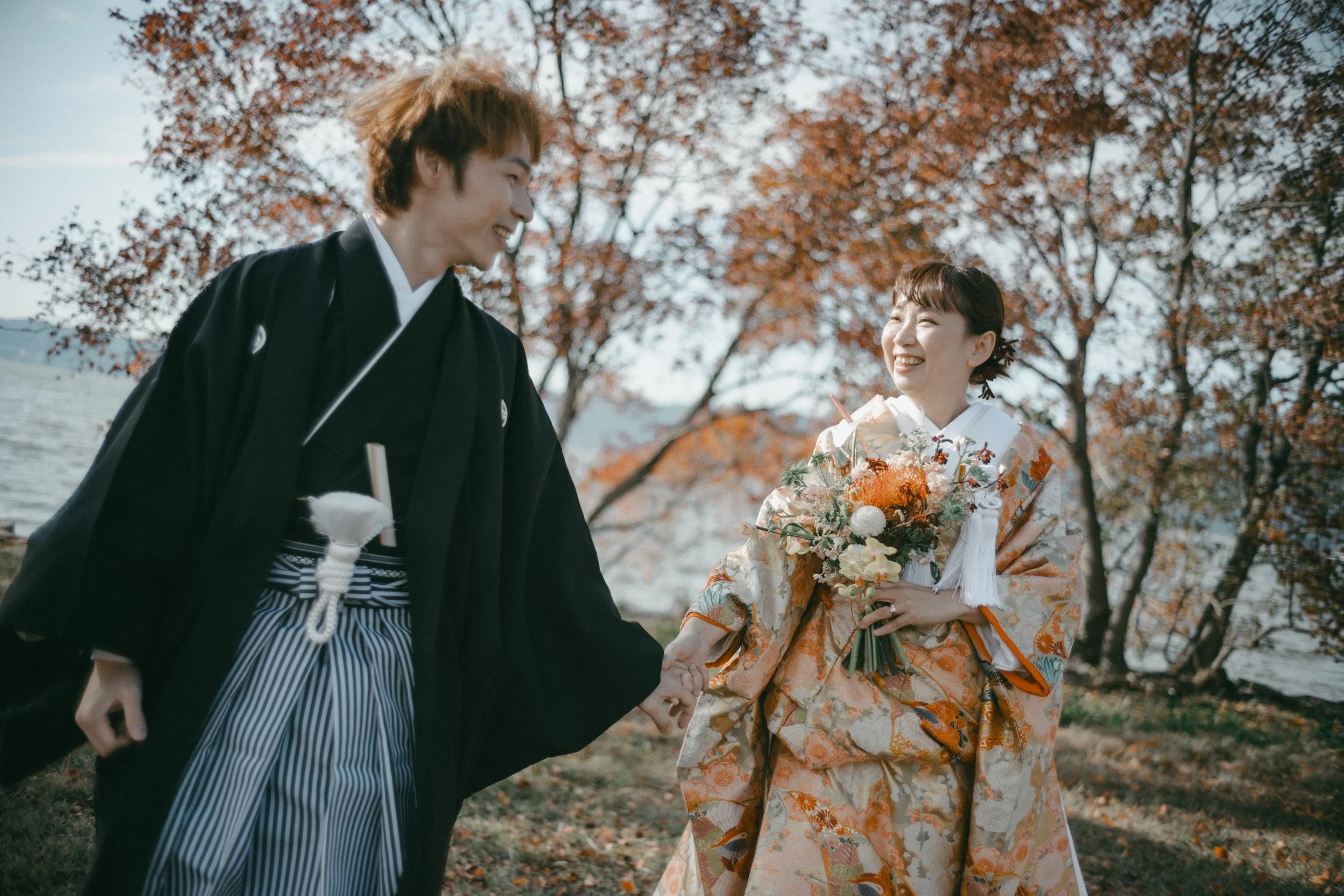 A happy couple in traditional Japanese wedding attire holding hands outdoors near a lake with autumn trees in the background.
