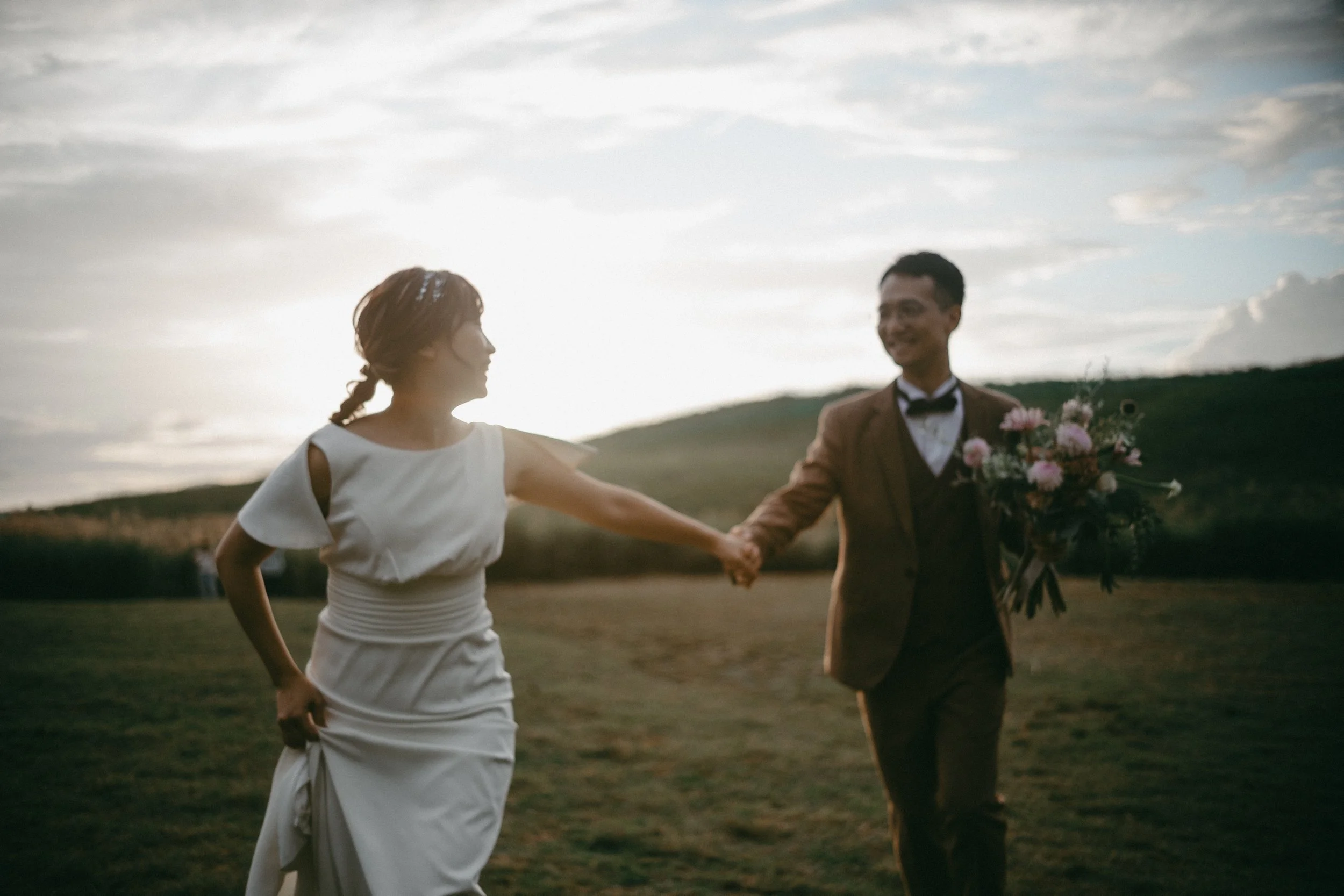 A newlywed couple holding hands and walking outdoors during sunset, with the bride in a white wedding dress and the groom in a brown suit holding a bouquet of flowers.