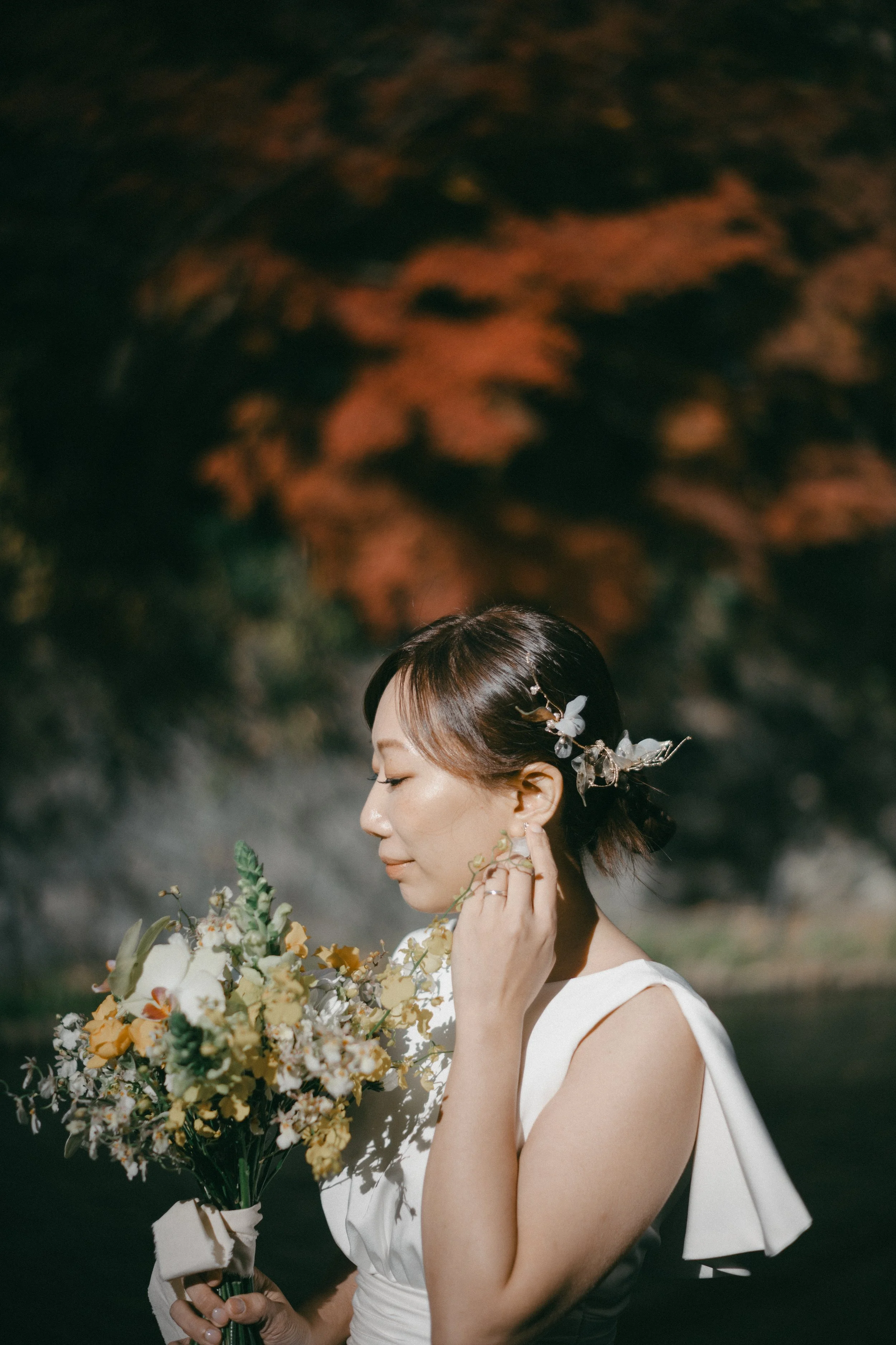 A woman in a white dress holding a bouquet of flowers, with her eyes closed and touching her ear, outdoors with autumn-colored trees in the background.