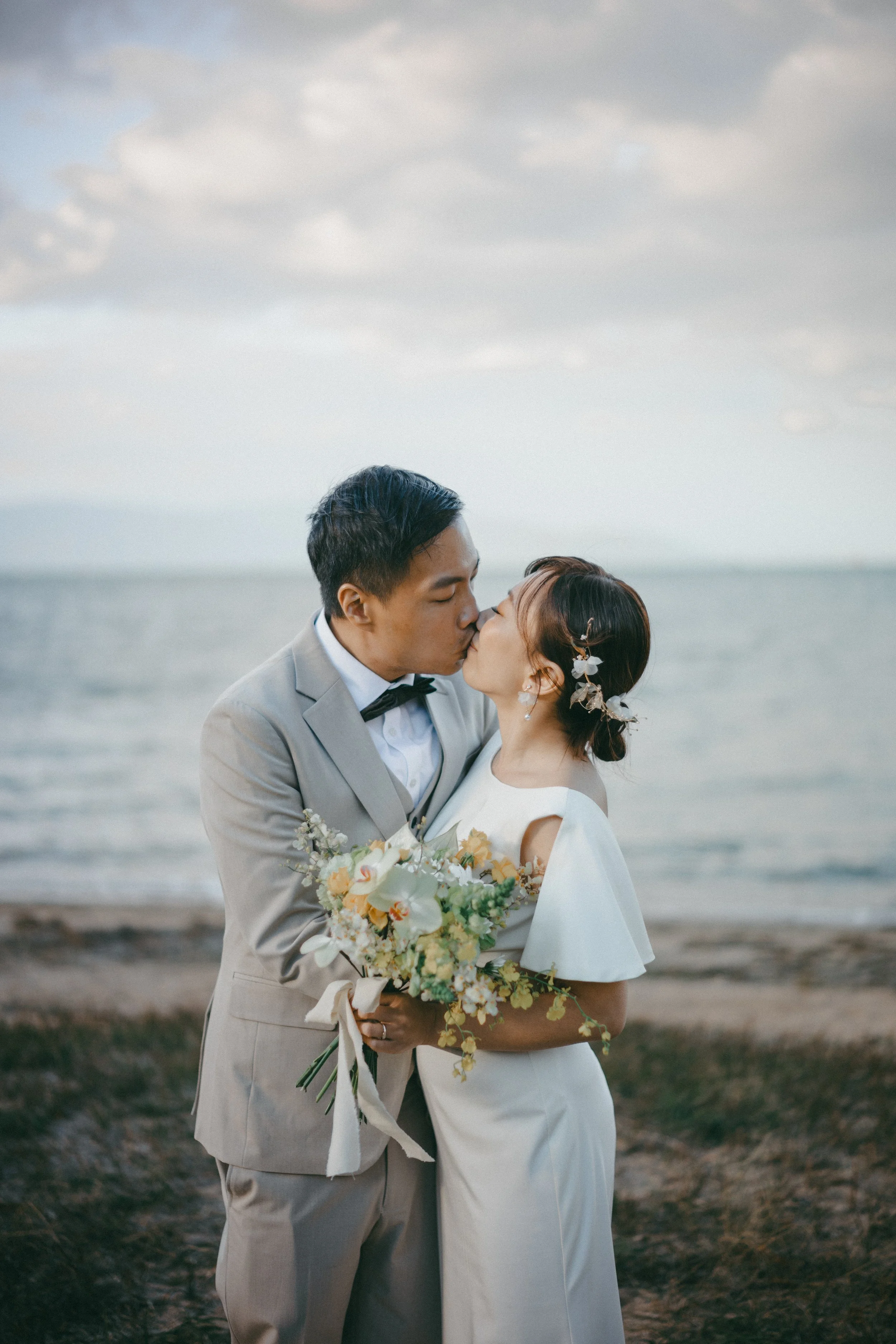 A couple in wedding attire sharing a kiss on a beach during sunset, with the ocean and cloudy sky in the background.