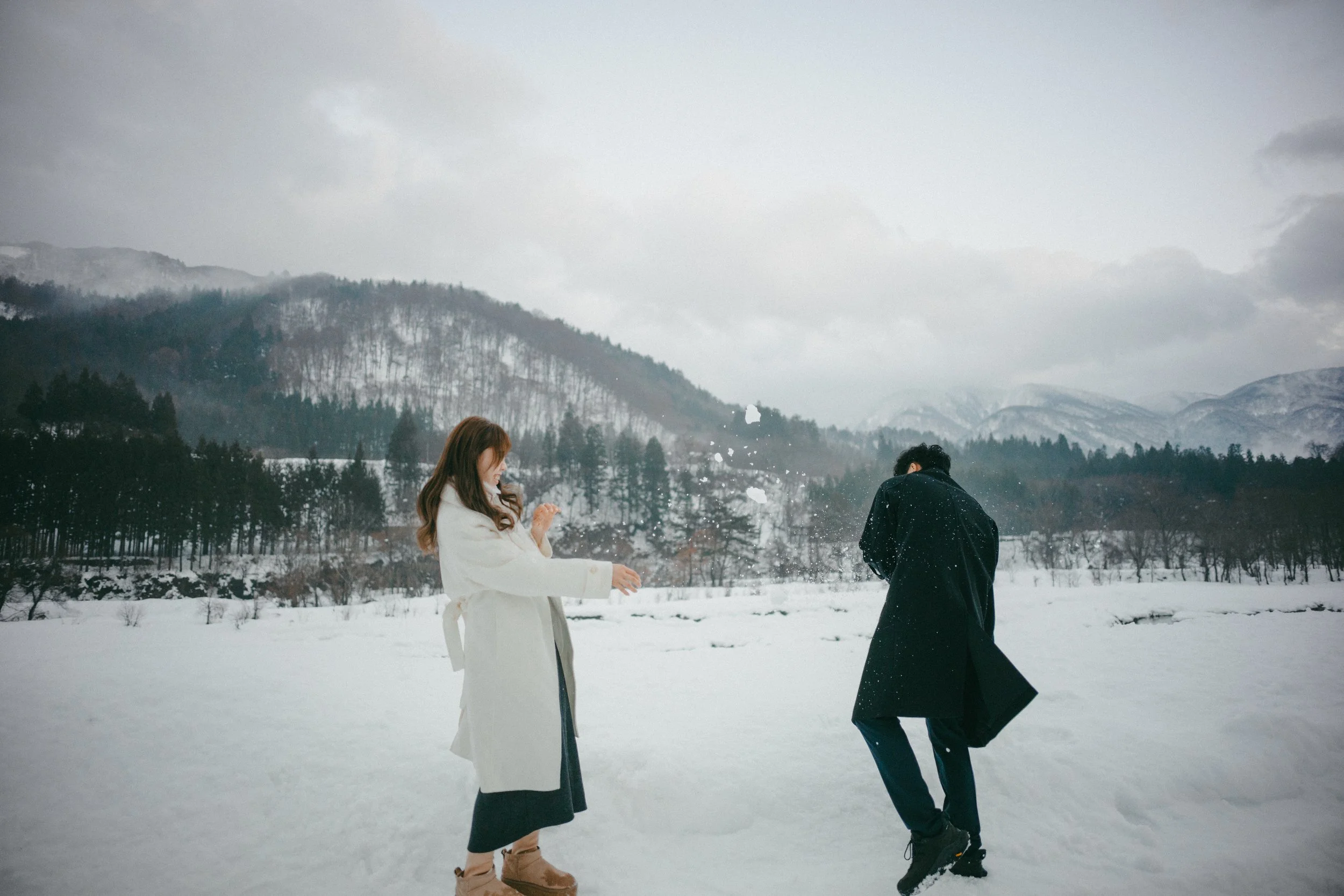 A woman and a man playing in the snow outdoors with a snowy mountain and forest in the background.