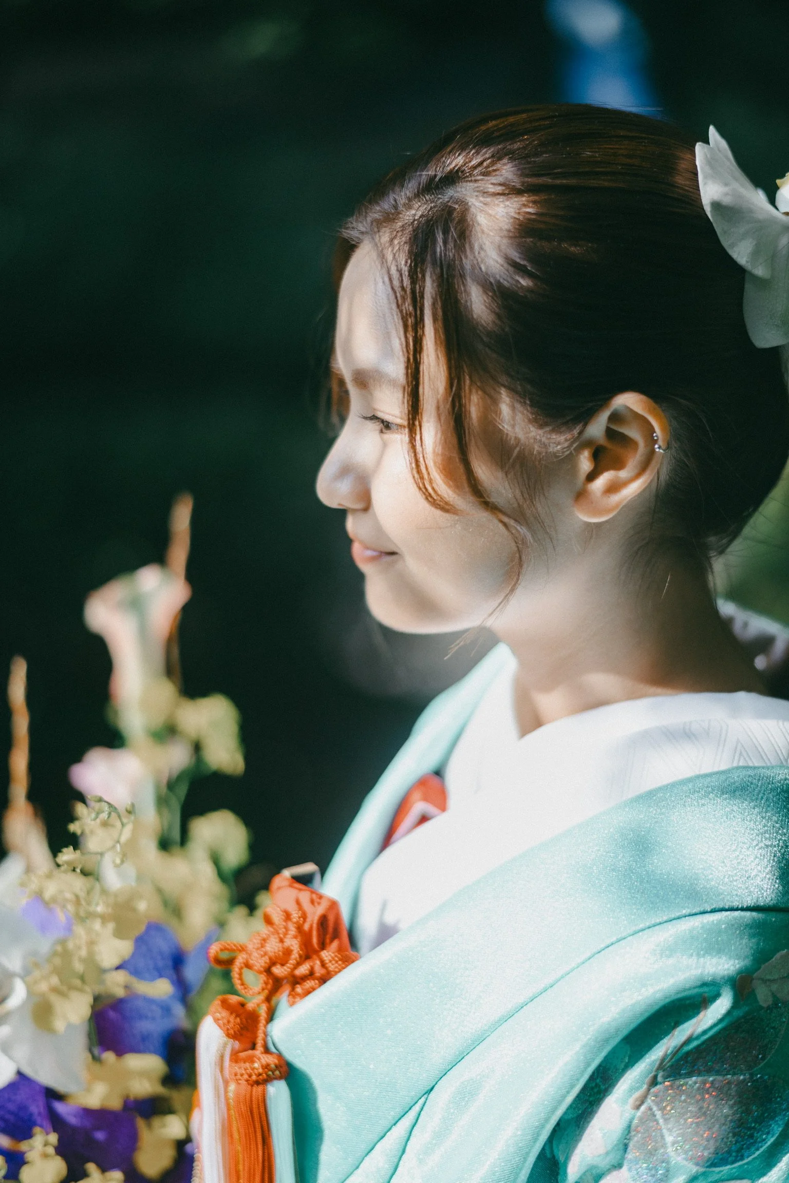 A young woman in traditional Japanese attire, holding a bouquet of flowers, with long hair styled in an elegant updo and a white flower accessory.