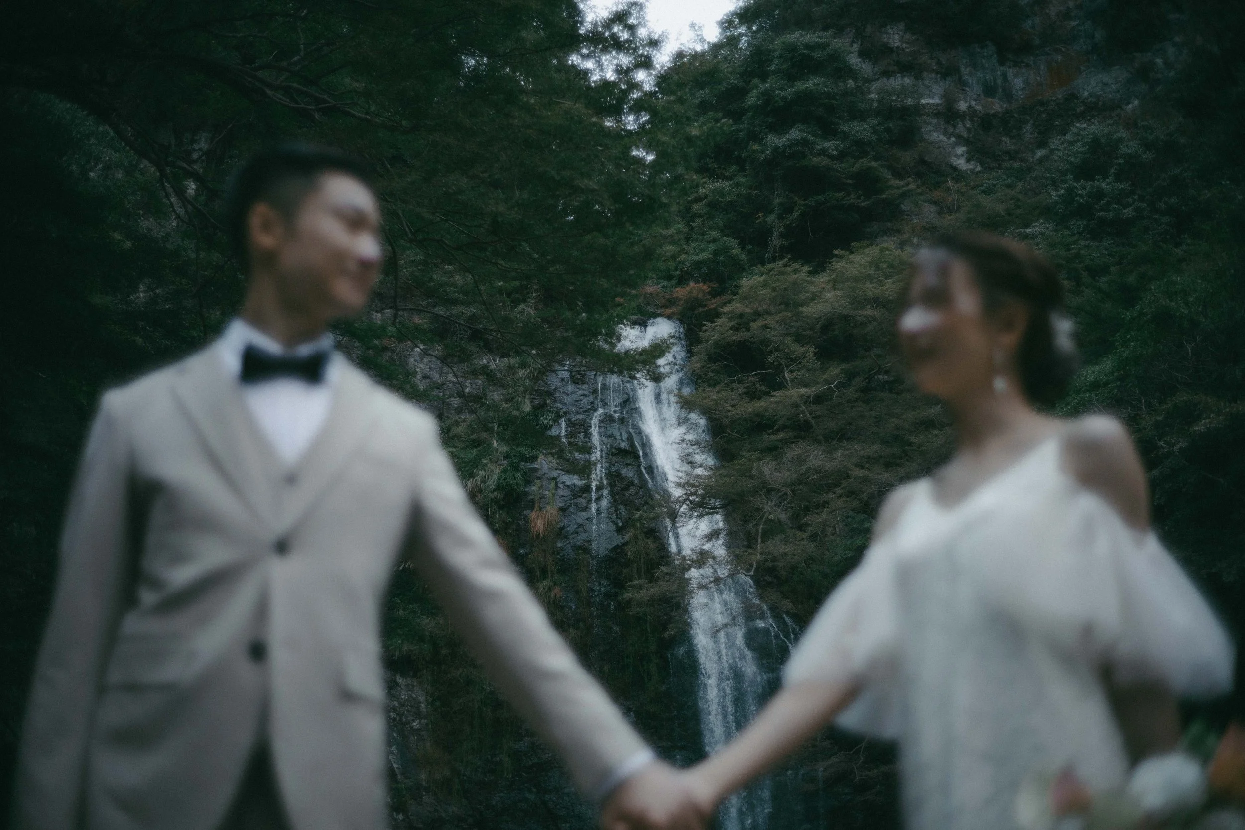 A couple dressed in wedding attire holding hands outdoors with a waterfall and forest in the background.
