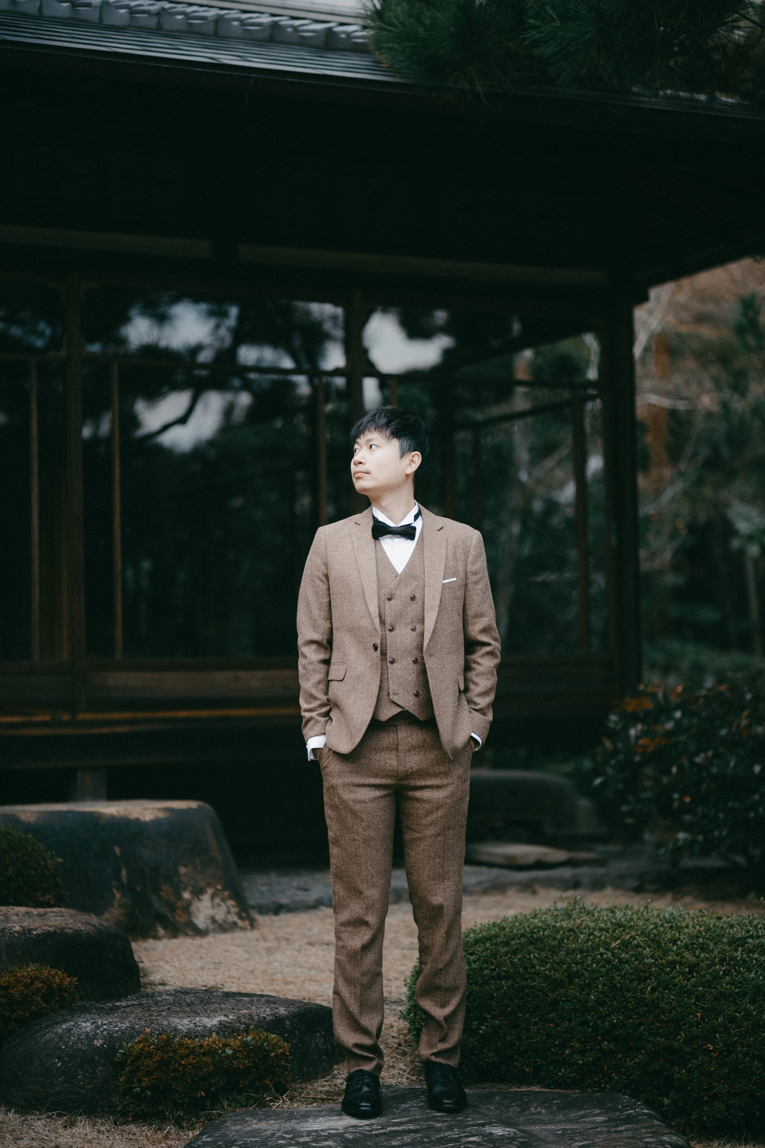 A young man in a brown suit with a bow tie and white shirt standing outdoors in front of a traditional Japanese structure surrounded by greenery.