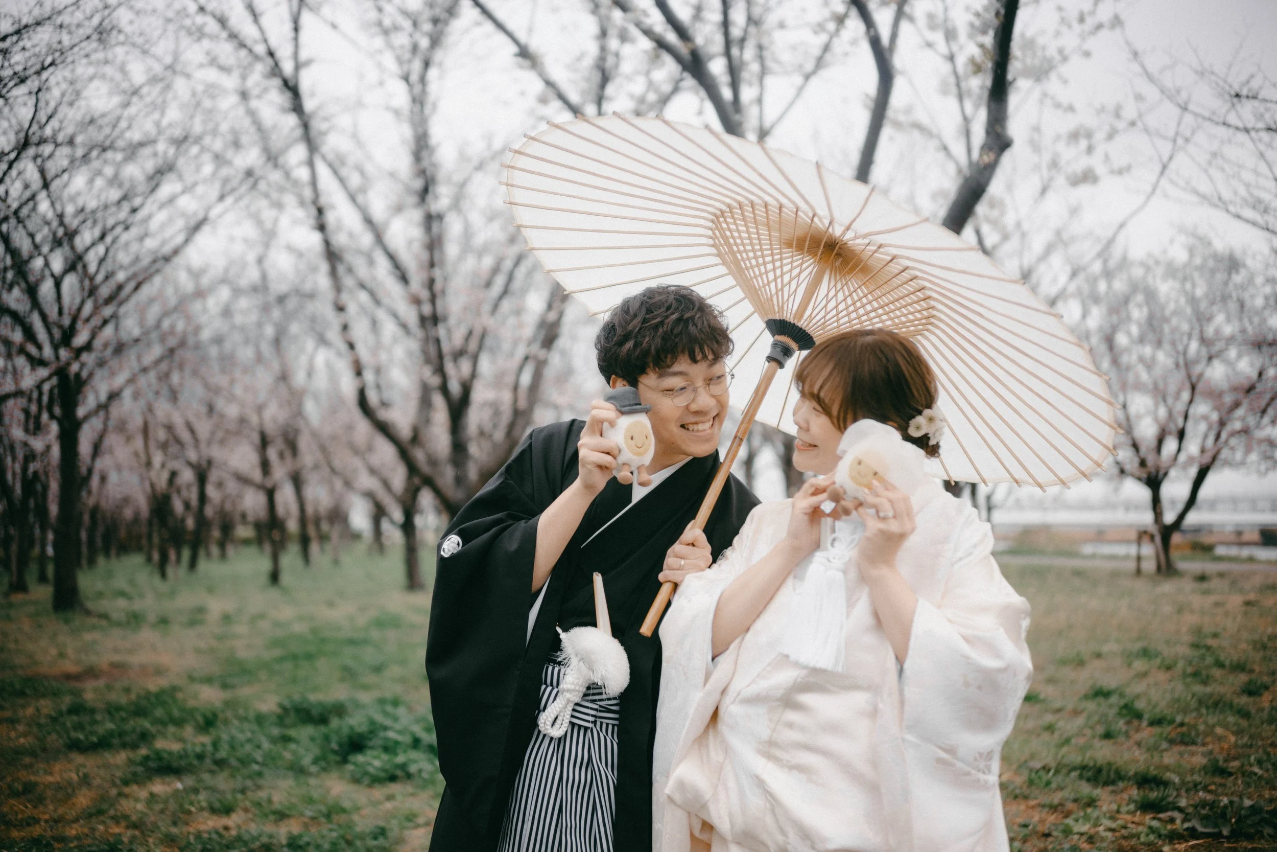 A couple dressed in traditional Japanese wedding attire standing in a park with cherry blossom trees. The man is holding a parasol and a small plush toy, while the woman is holding a plush toy. They are smiling at each other, sharing a joyful moment.