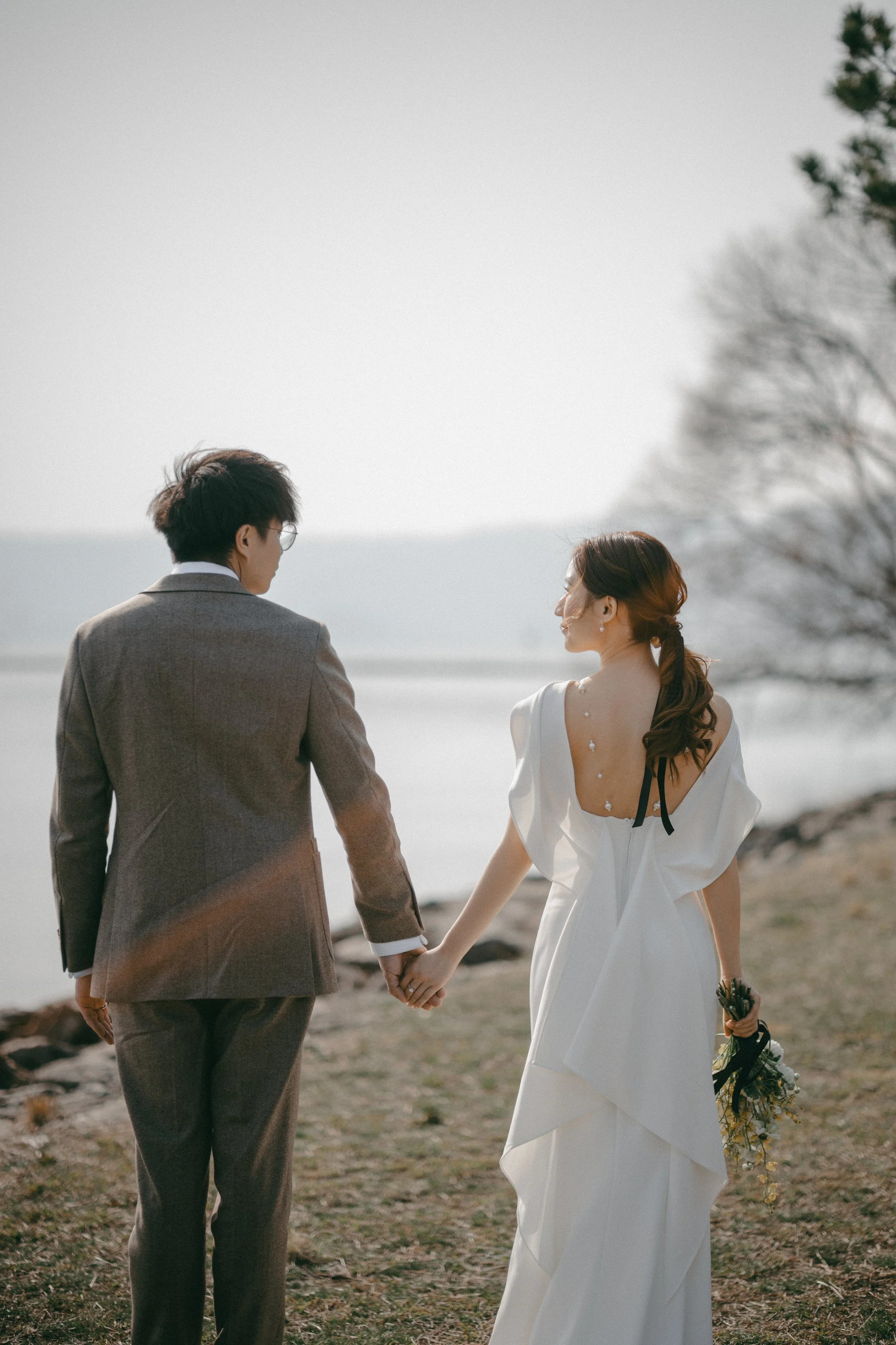A couple holding hands on a beach near the water, with trees in the background, during daytime.