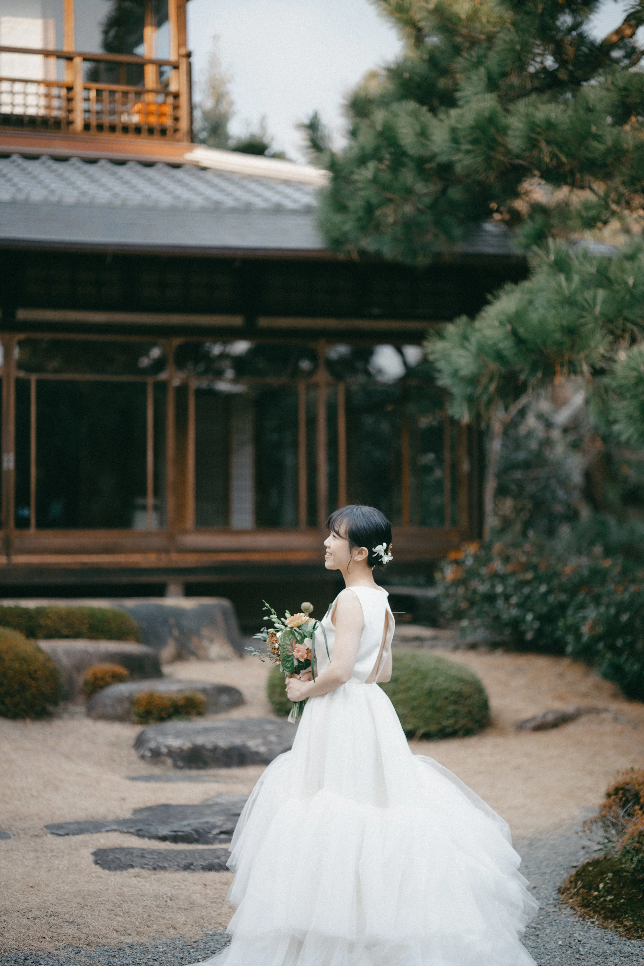 A bride in a white wedding dress holding a bouquet in a traditional Japanese garden with a wooden house in the background.