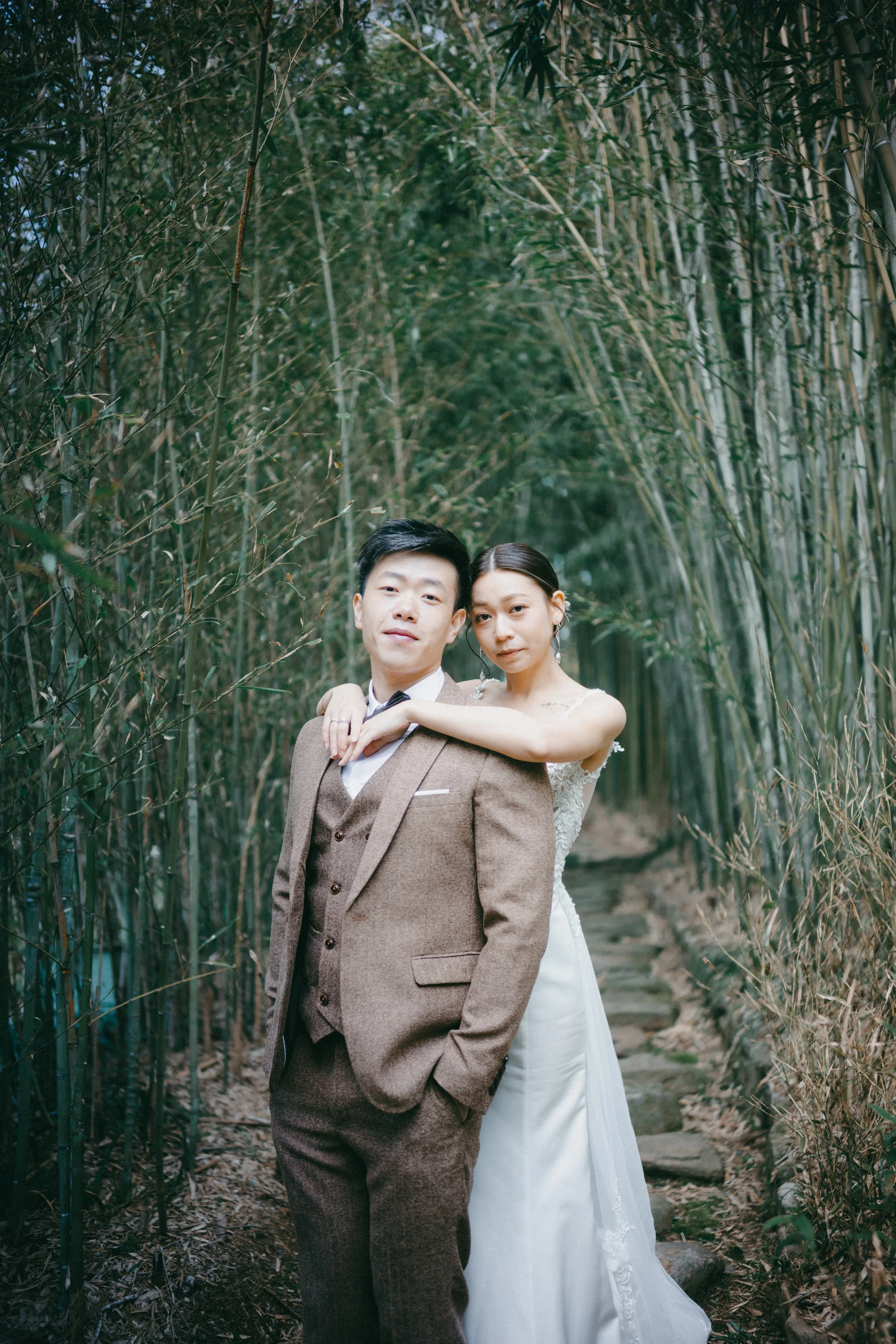 A bride and groom stand close together in a bamboo forest, with the bride resting her arm on the groom's shoulder.