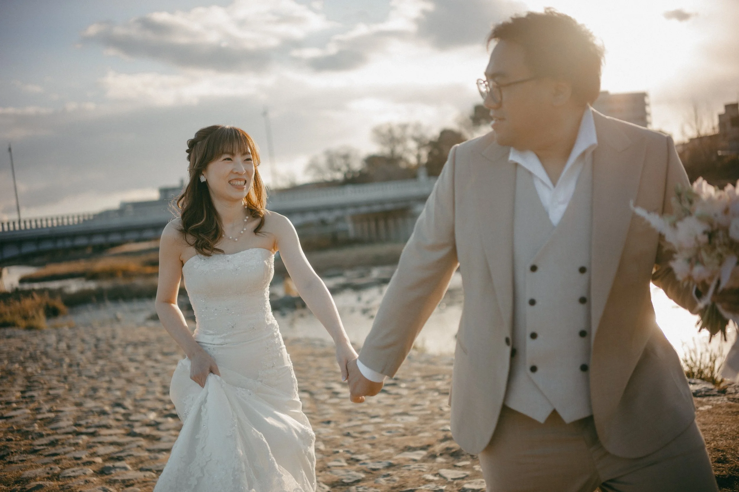 A couple in wedding attire holding hands outdoors during sunset, smiling and laughing, with a bridge and water in the background.
