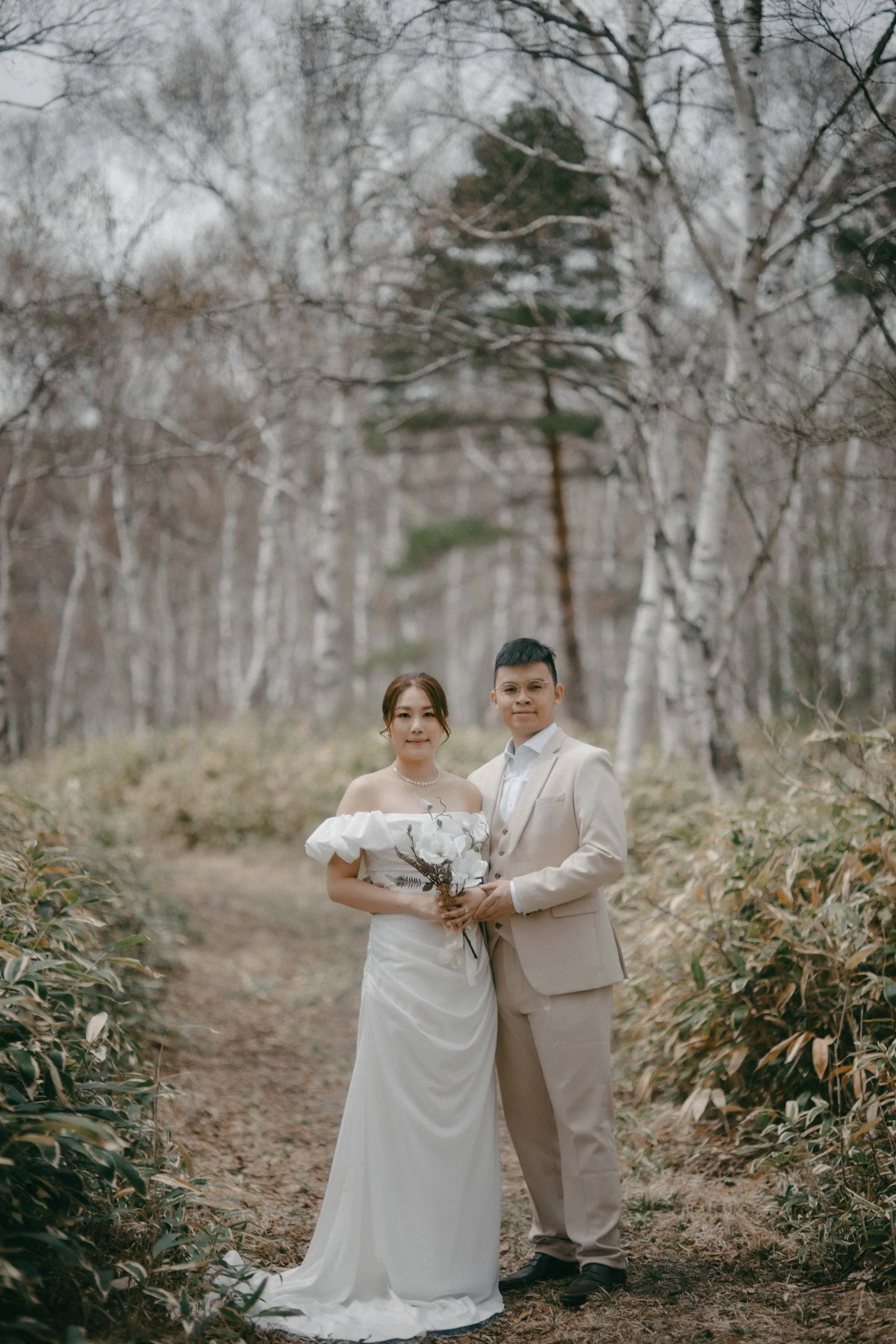 A bride and groom standing on a dirt path in a forest, dressed in wedding attire, holding a bouquet of white flowers and branches.