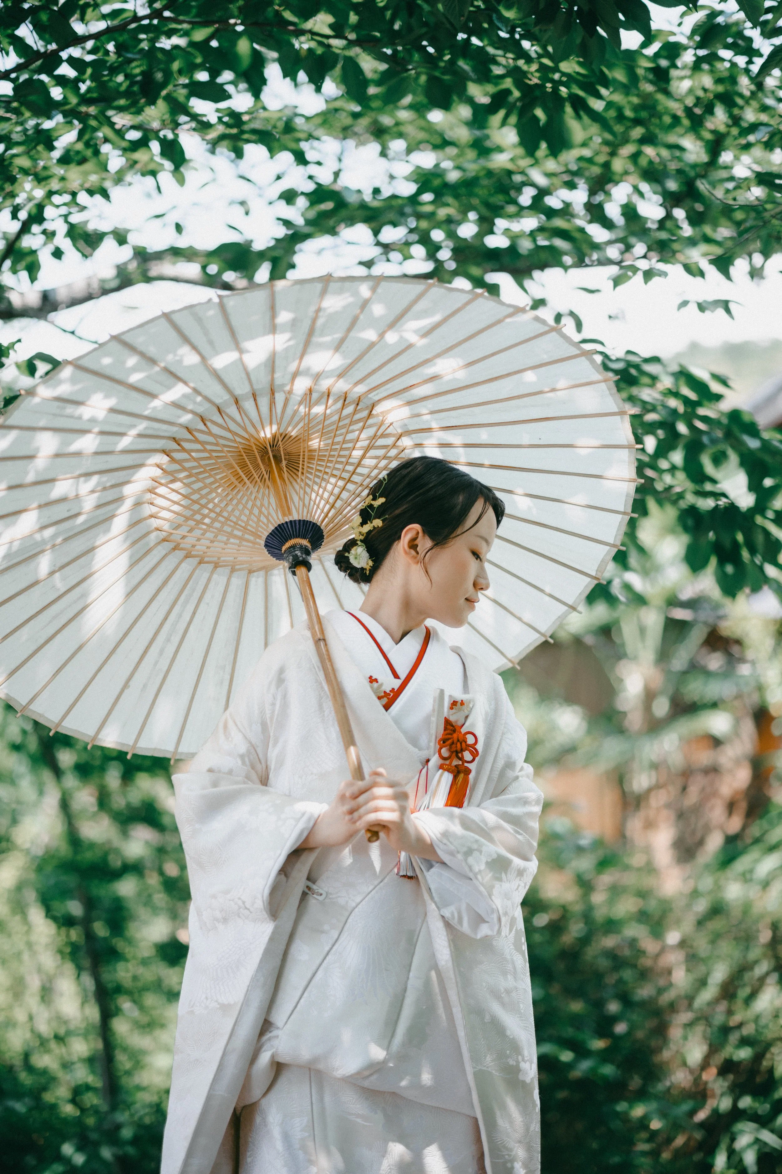 A woman dressed in traditional white Japanese bridal kimono holding a white parasol, standing outdoors among green foliage.