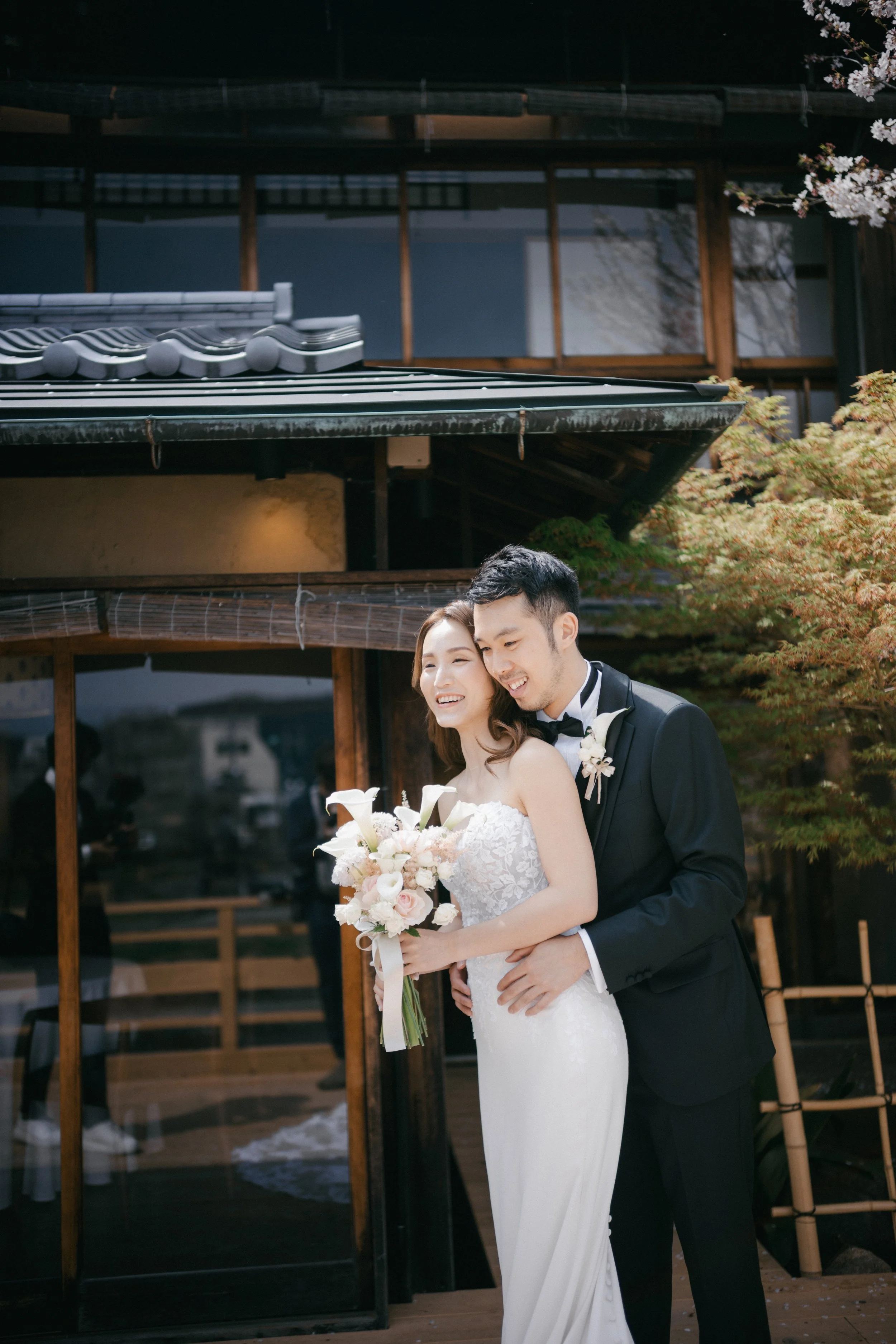 A bride and groom standing outside a traditional Japanese building, smiling and holding a bouquet of white flowers, during their wedding celebration.