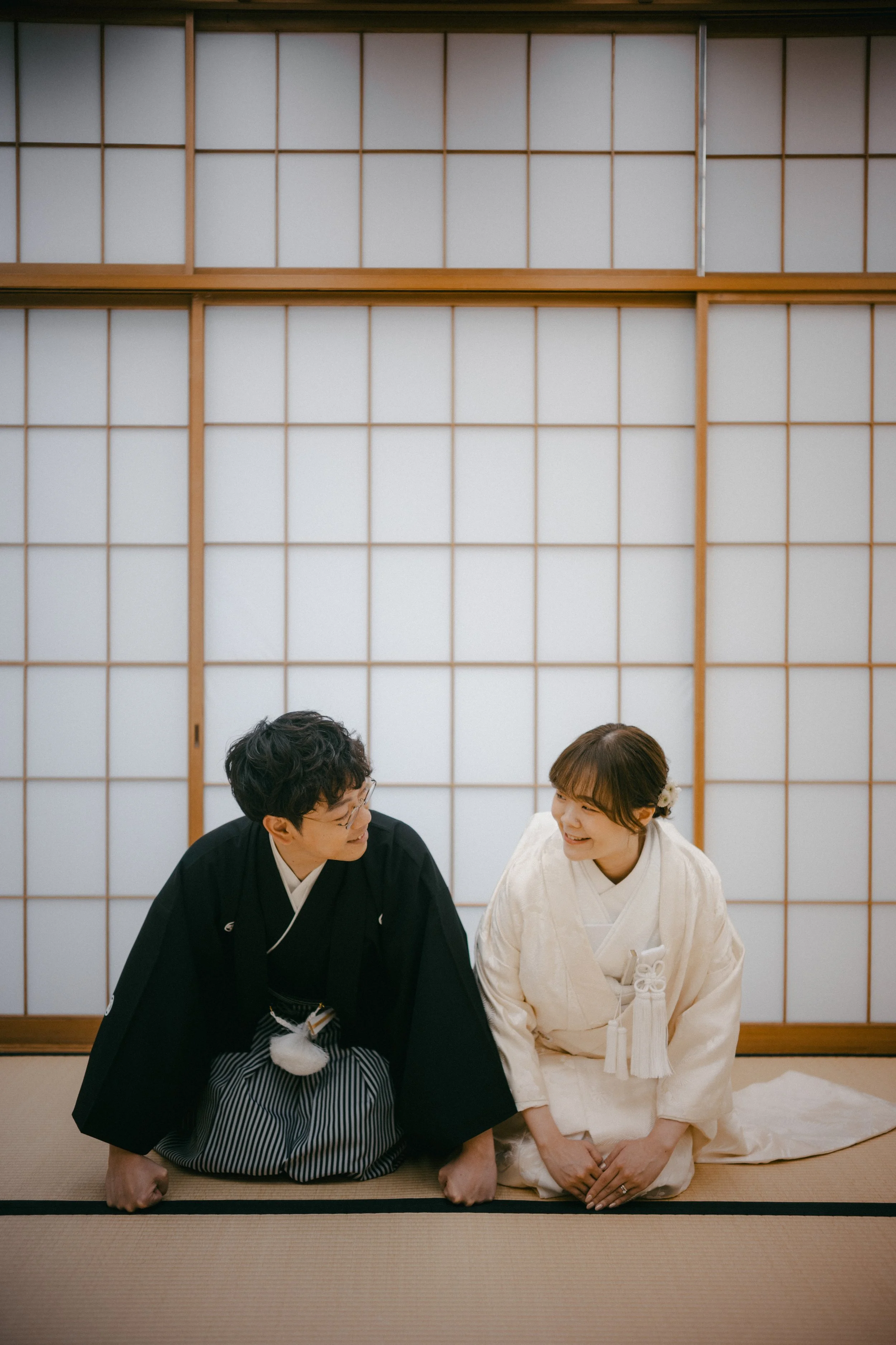 A man and woman dressed in traditional Japanese attire, sitting on tatami mats in front of shoji screens, smiling and looking at each other.