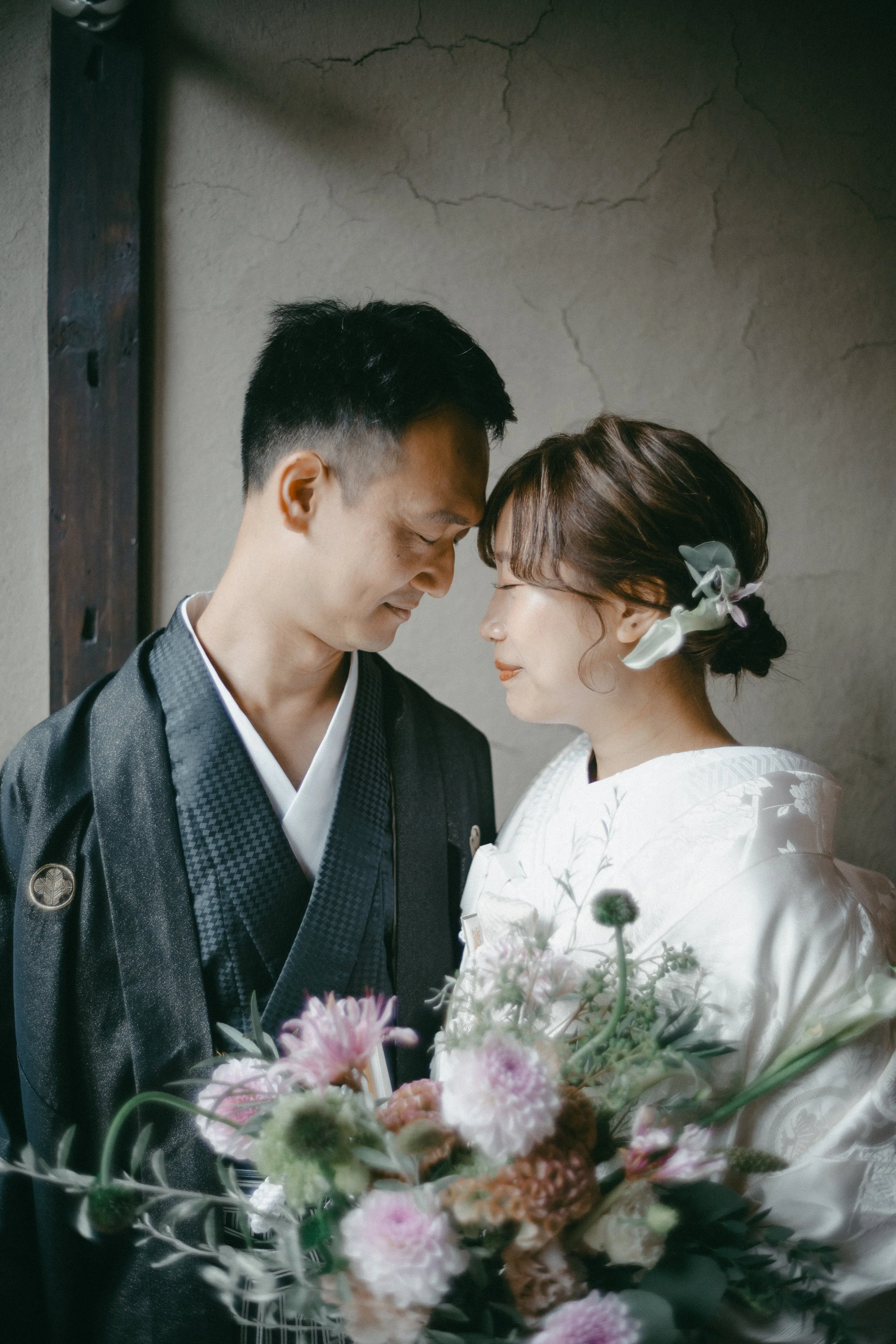 A couple dressed in traditional Japanese wedding attire sharing a tender moment, with foreheads touching, in front of a textured wall, while holding a bouquet of pink, white, and green flowers.