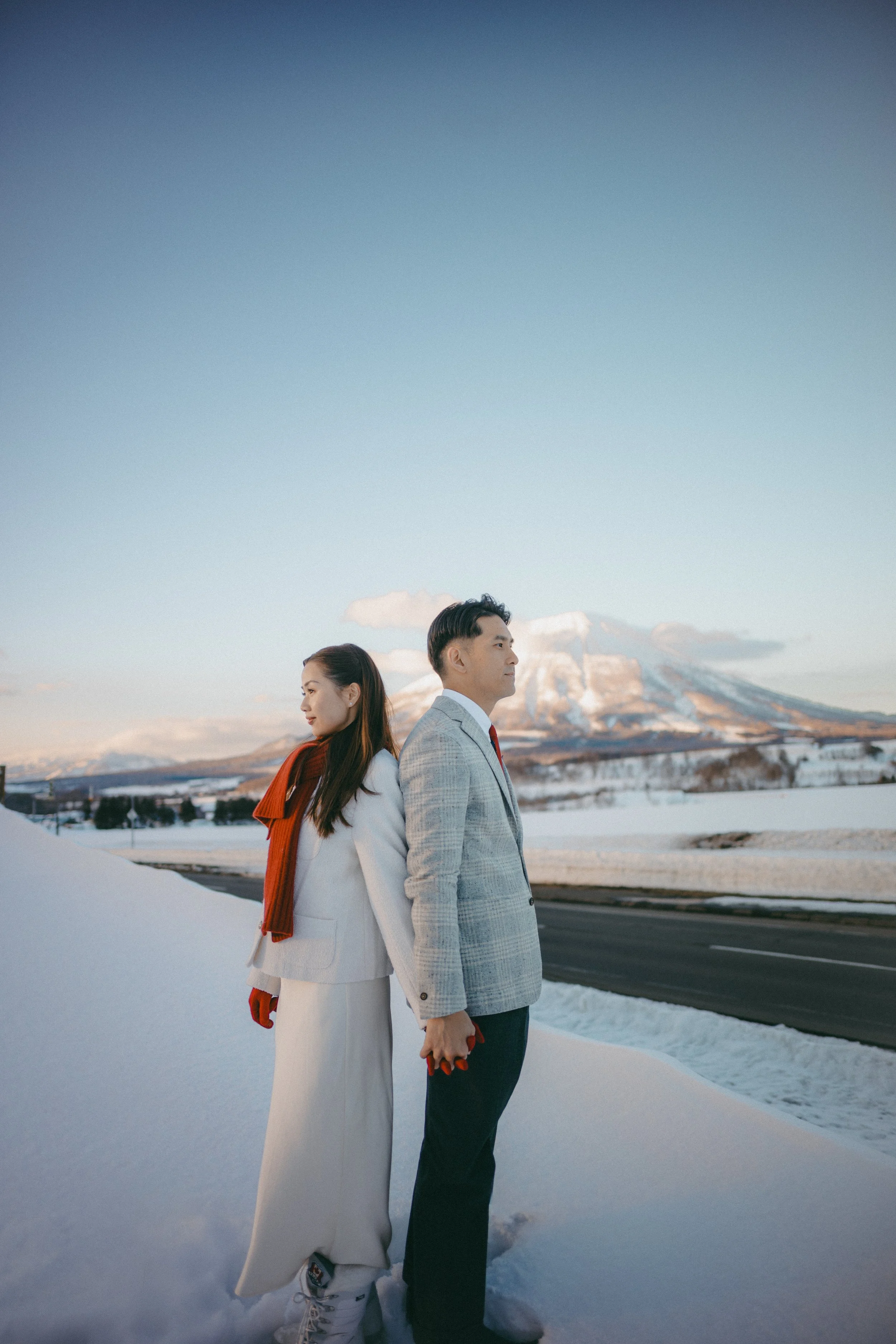 A man and woman in formal winter clothing standing back to back outdoors with snowy landscape and mountain in the background.